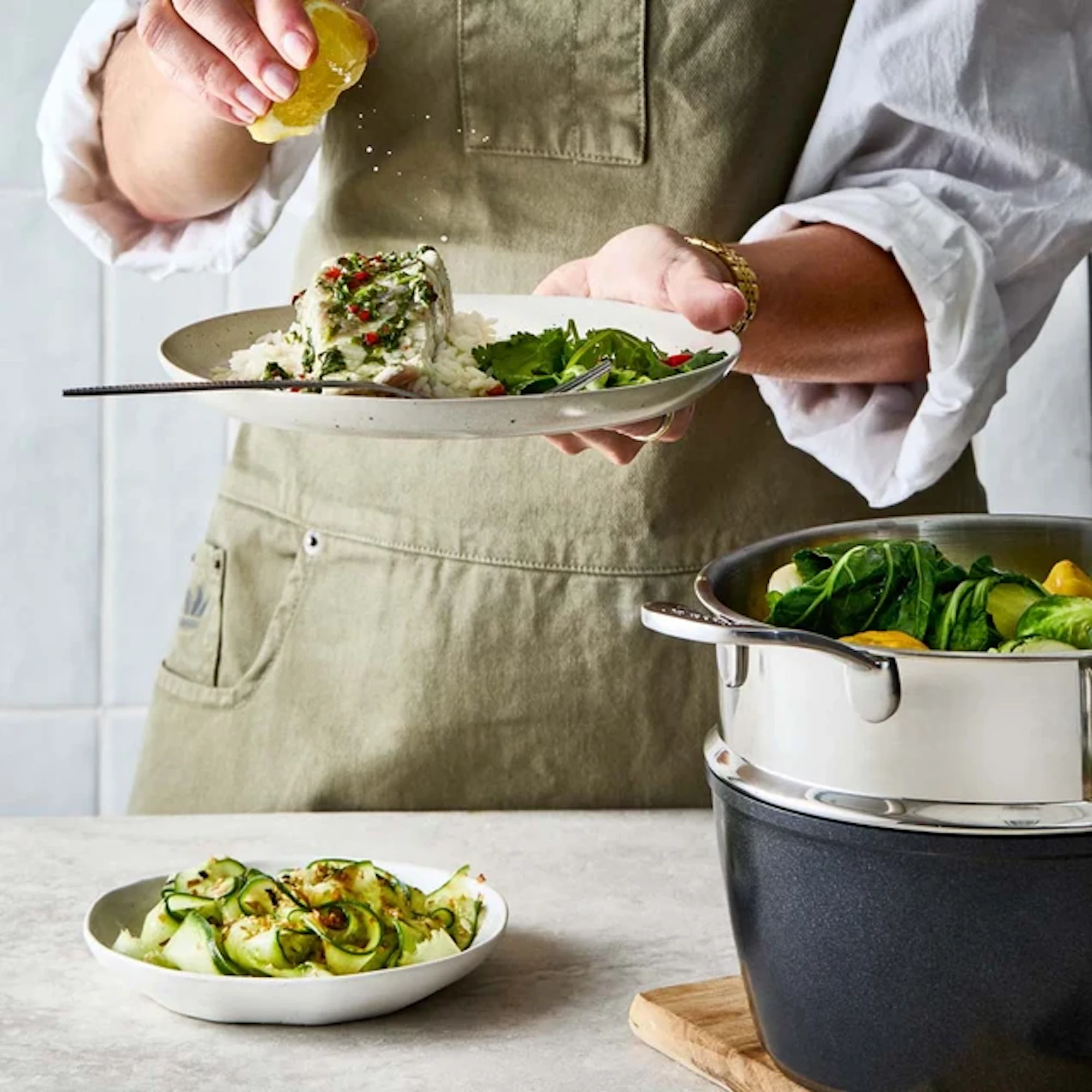 A person is holding a plate of fish and rice while squeezing a lemon over it. In the foreground, a bowl of sliced zucchini is set next to a stacked metal pot filled with vegetables.
