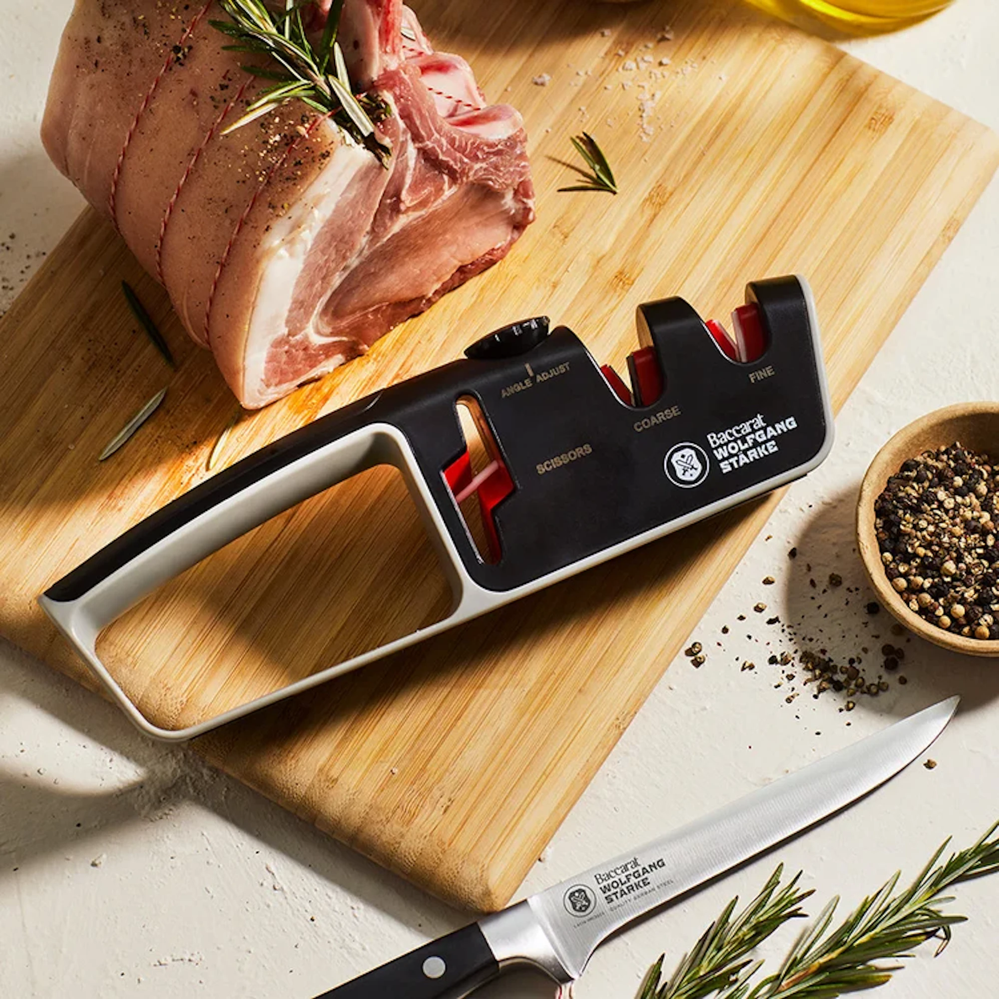 A Baccarat Wolfgang Starke knife sharpener rests on a wooden cutting board beside a raw rolled roast, a bowl of peppercorns, and a Baccarat Wolfgang Starke knife.