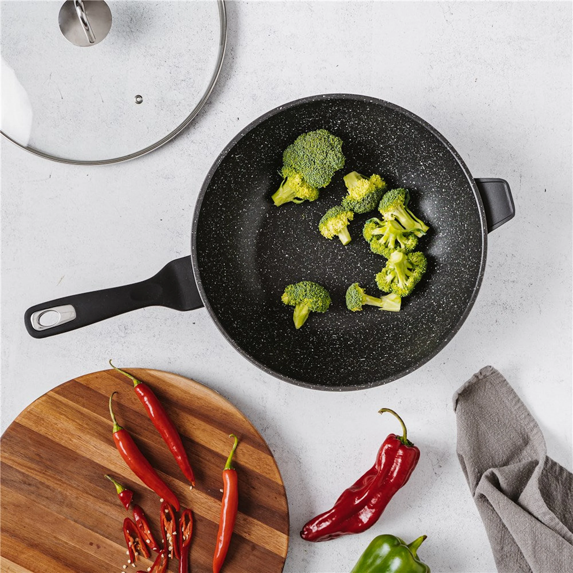 Broccoli florets are arranged in a black frying pan on a light gray stone background, with red chilies on a wooden board, peppers, a glass lid, and a gray cloth nearby.