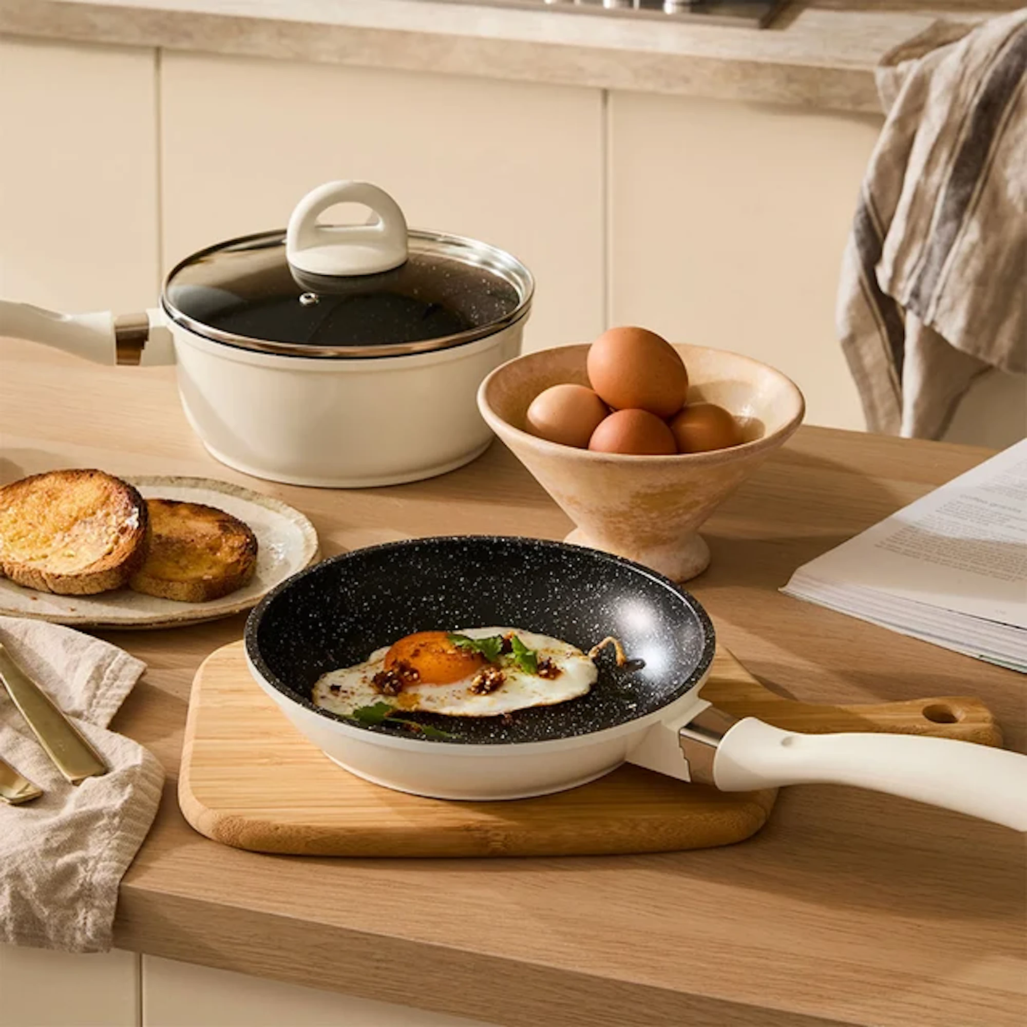 A fried egg is in a pan on a wooden board, with a pot, toast, and a bowl of eggs nearby.