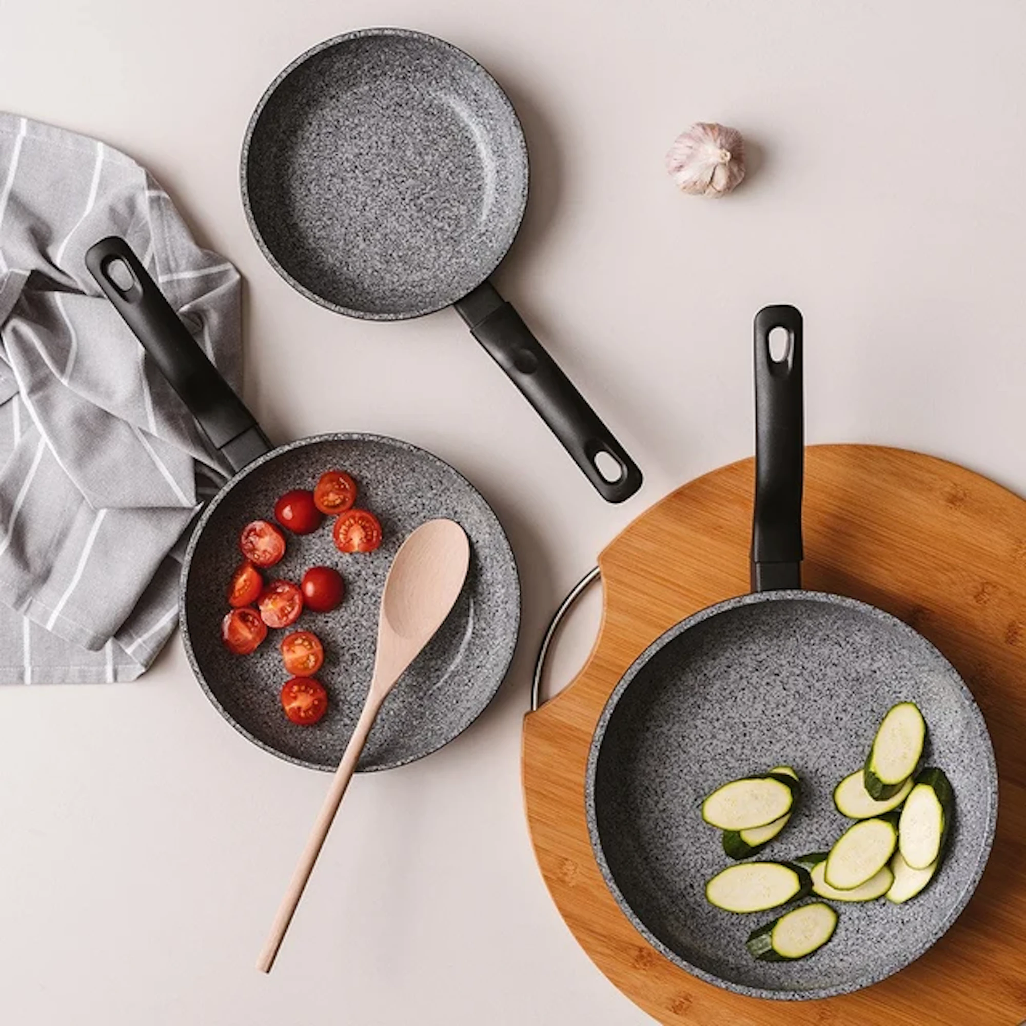 Three frying pans are arranged on a light surface with sliced tomatoes, zucchini, and a wooden spoon.