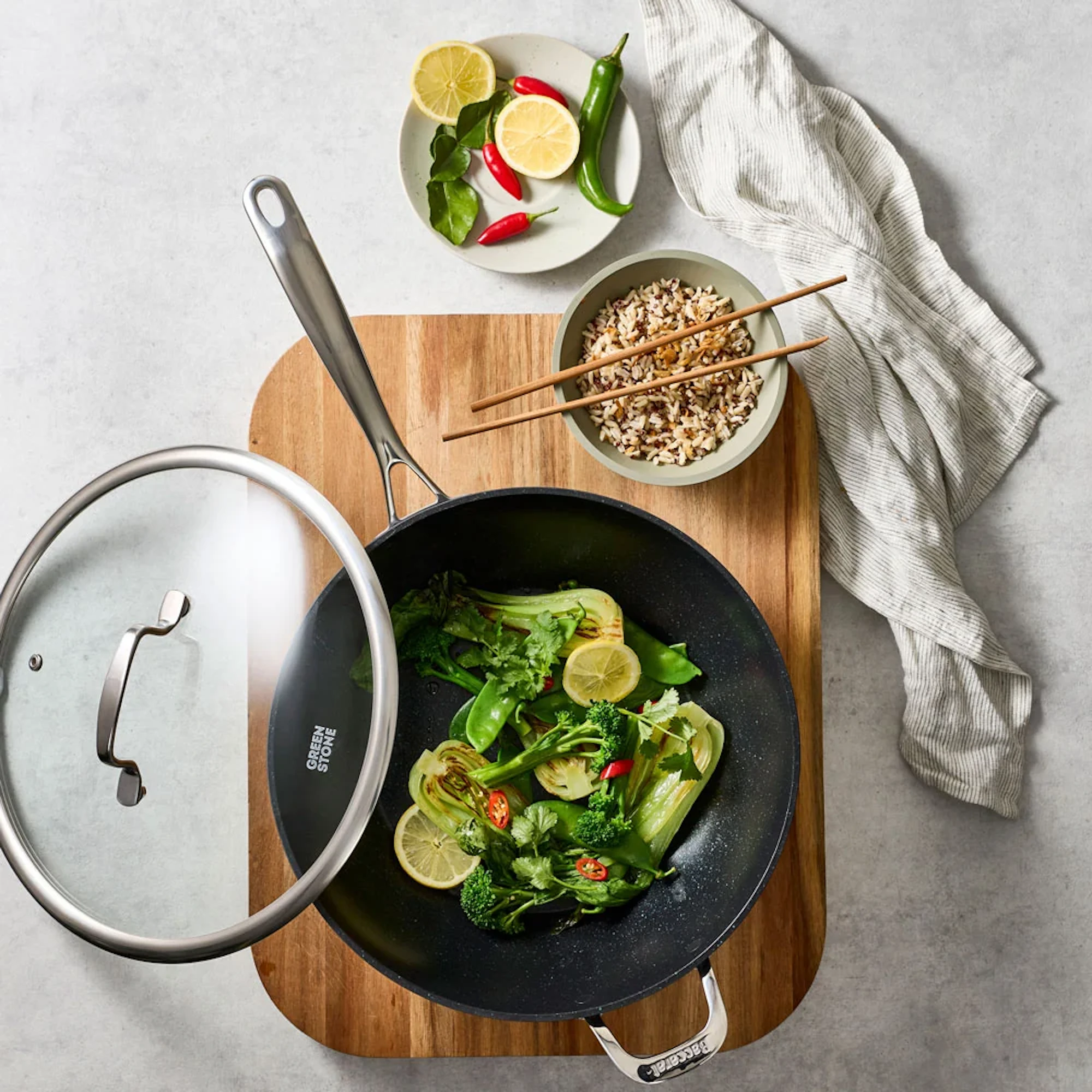 Green vegetables with lemon slices and chili are cooked in a black wok on a wooden board, with rice, fresh ingredients, and a striped cloth on a light gray stone background.