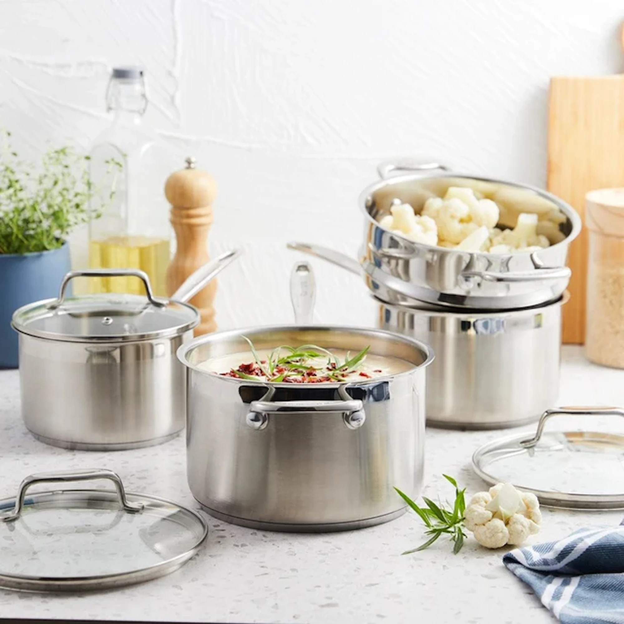 A collection of stainless steel cookware is arranged on a kitchen counter. Several pots, glass lids, and a stack with cauliflower are set against a white wall background.