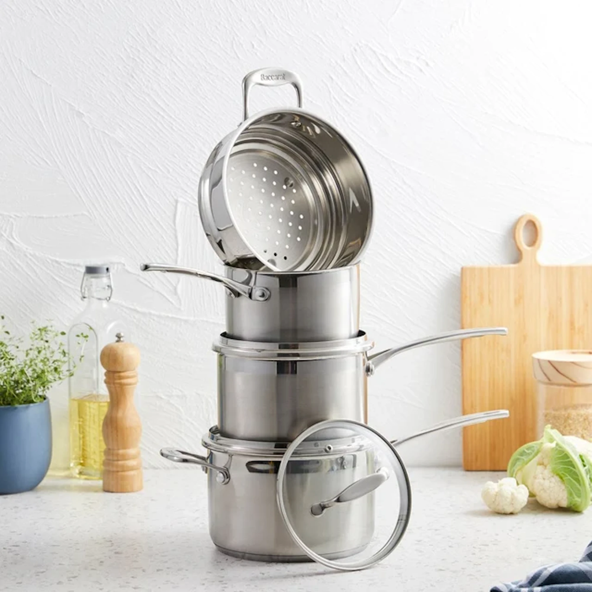 A tall stack of stainless steel pots, pans, and a steamer basket is balanced on a kitchen counter against a white wall background