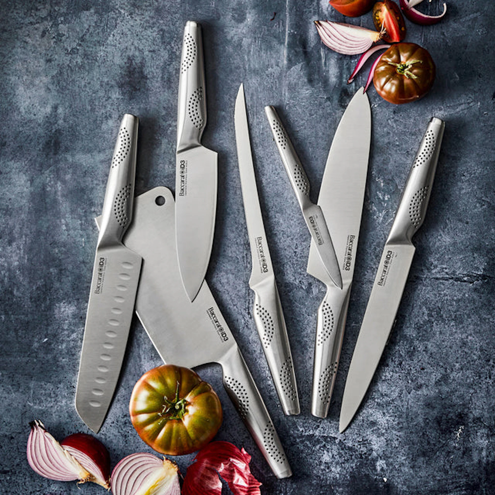 Several kitchen knives are arranged on a dark surface alongside cut vegetables.