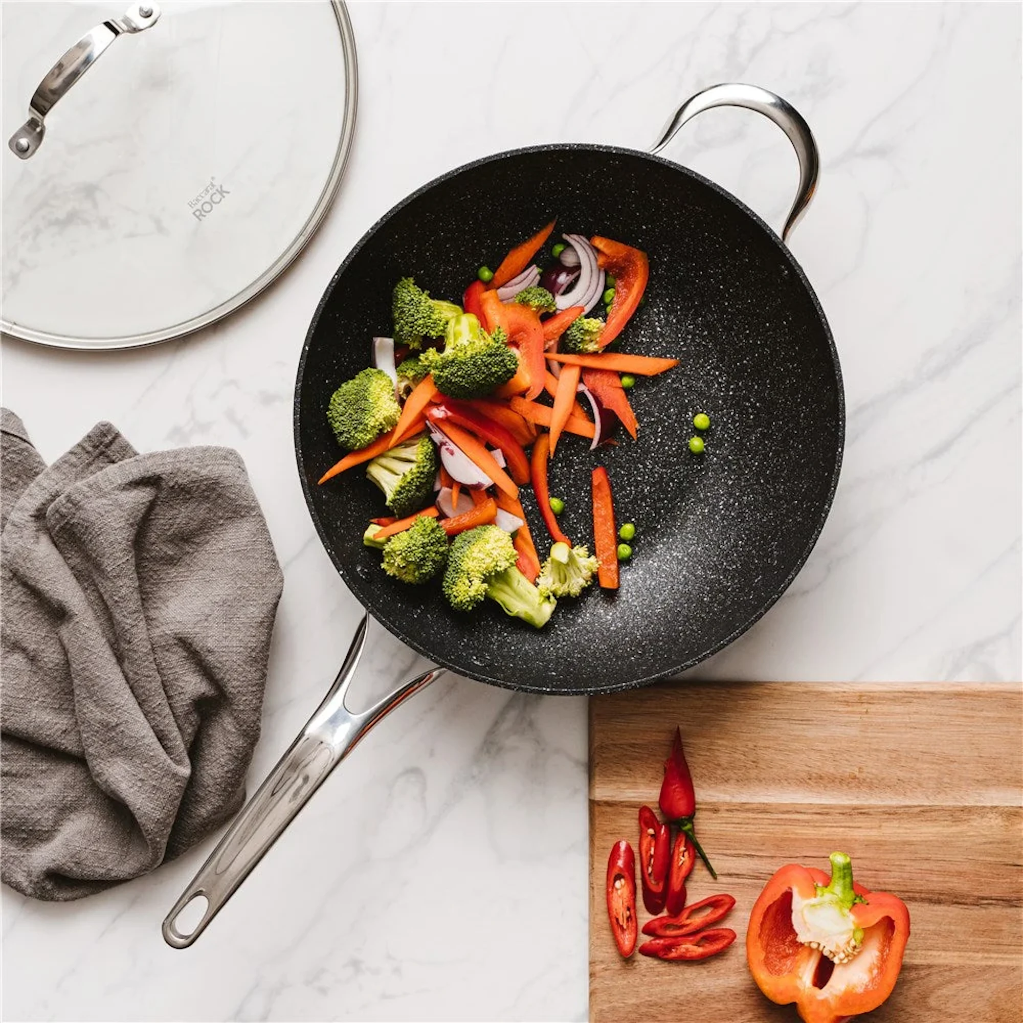 Broccoli, carrot strips, red pepper, and onion are arranged in a black wok on a white marble background, with a wooden board, sliced chili, and a gray cloth nearby.