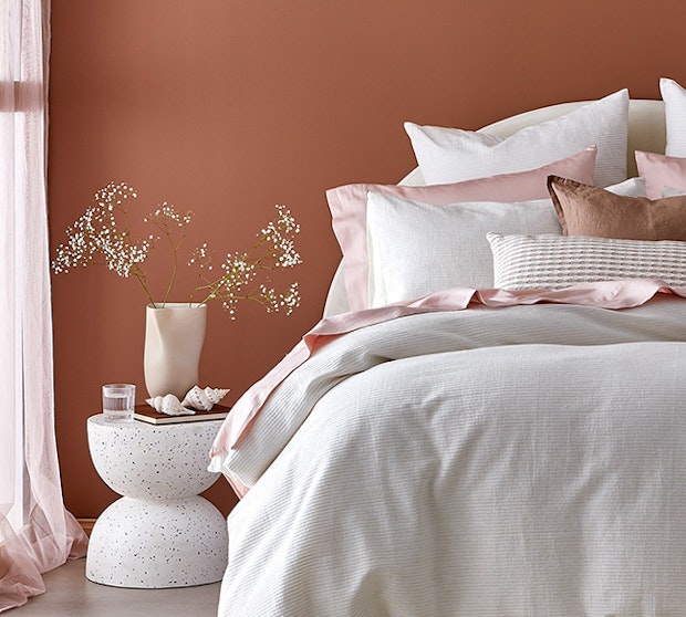 Home Beautiful Collection. Bedroom styling with white quilt cover and pink sheets, layered with cushions, with a side table and flowers