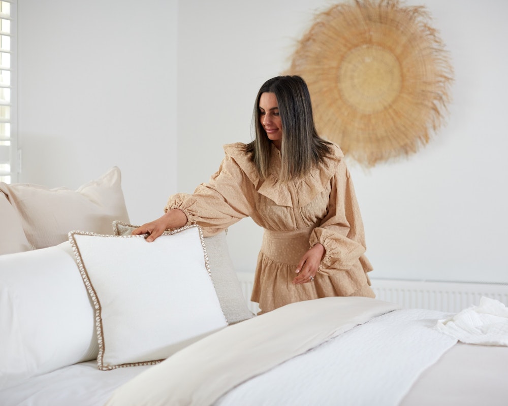 Woman fluffing a cushion on a neutral based bed in a light room. Home Beautiful collection.