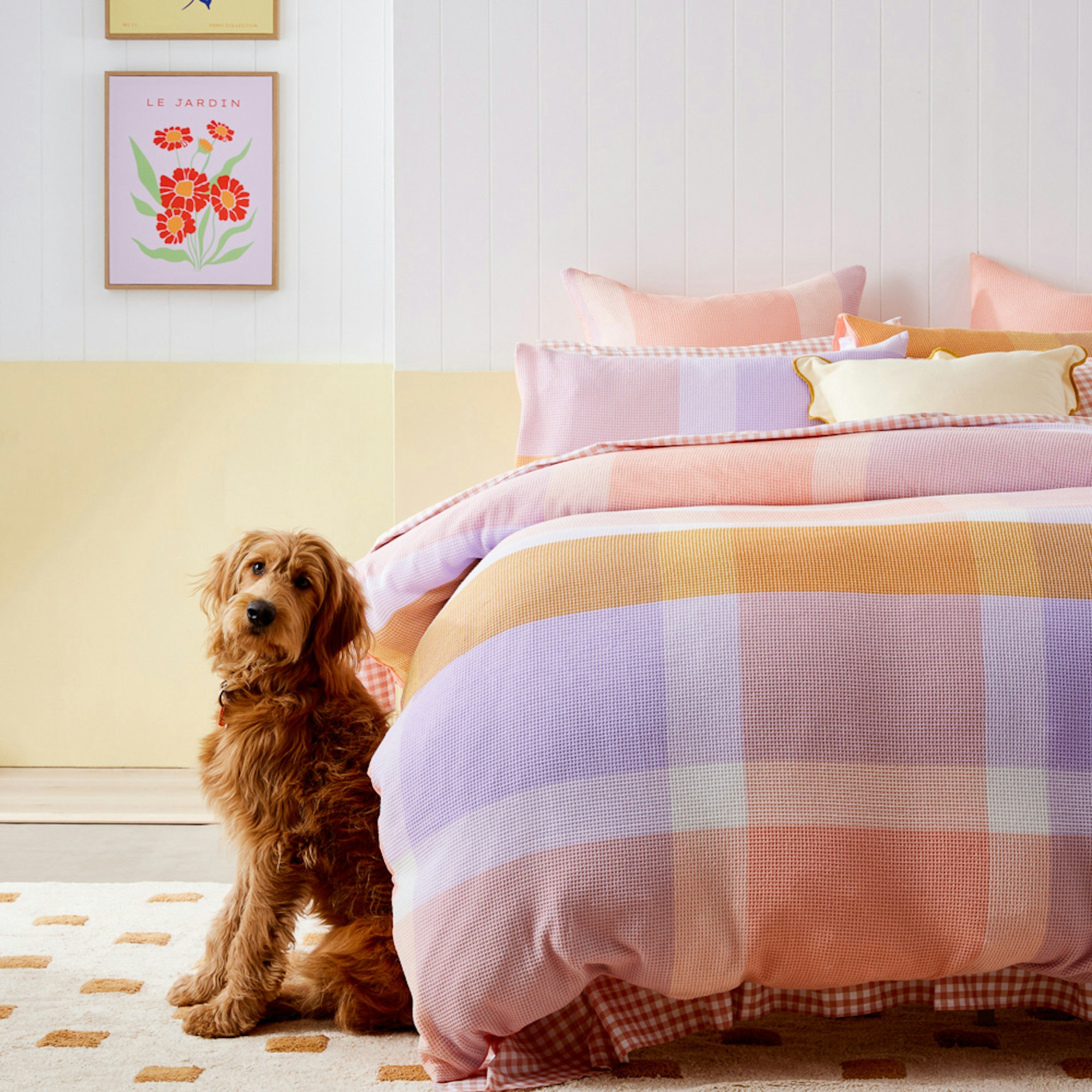 Pastel coloured gingham quilt cover set layered with cushion and brown dog sitting by the side of the bed.