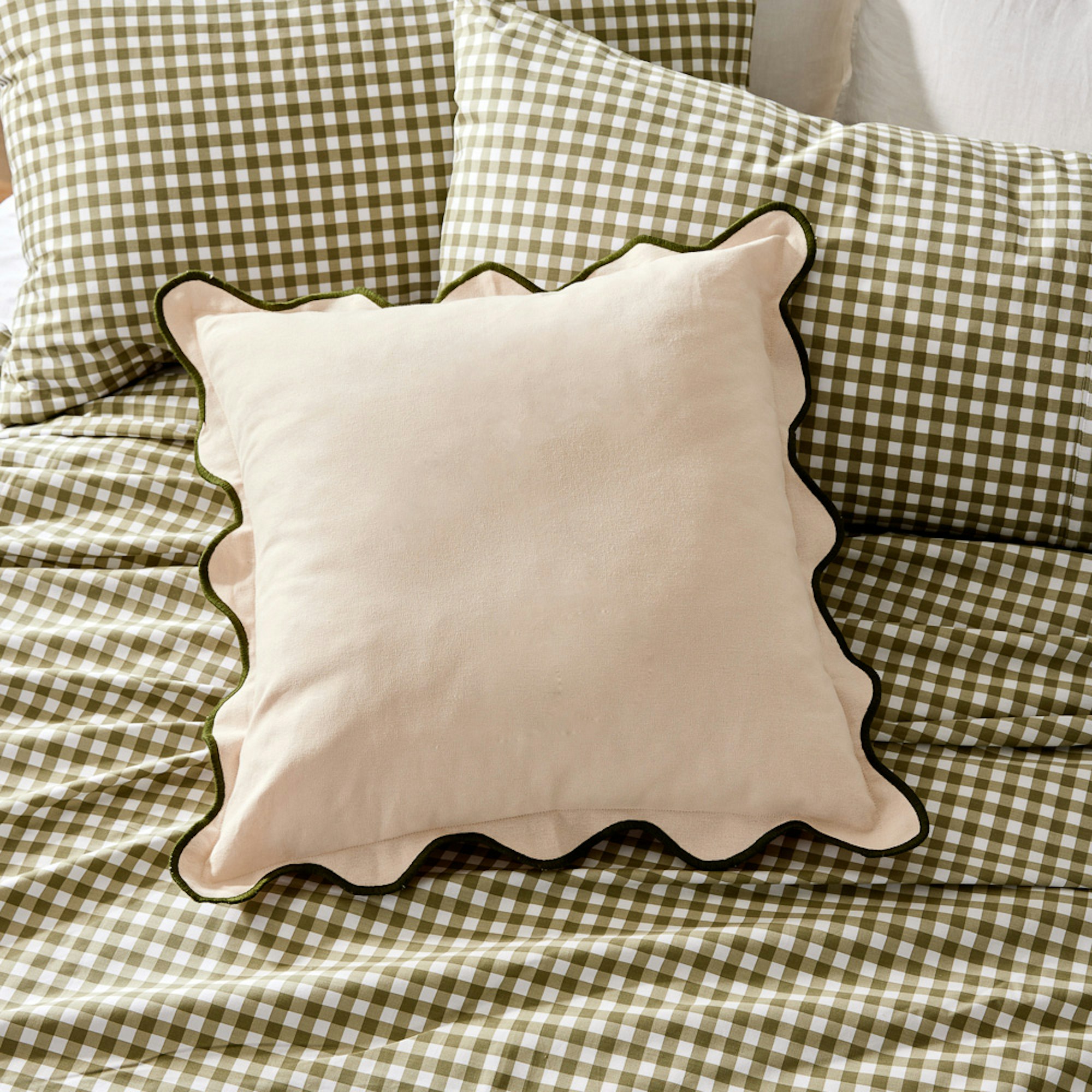MyHouse SS23 collection. Lola cushion. scallop edged cushion in natural sitting on top of bed with green and white gingham sheets.