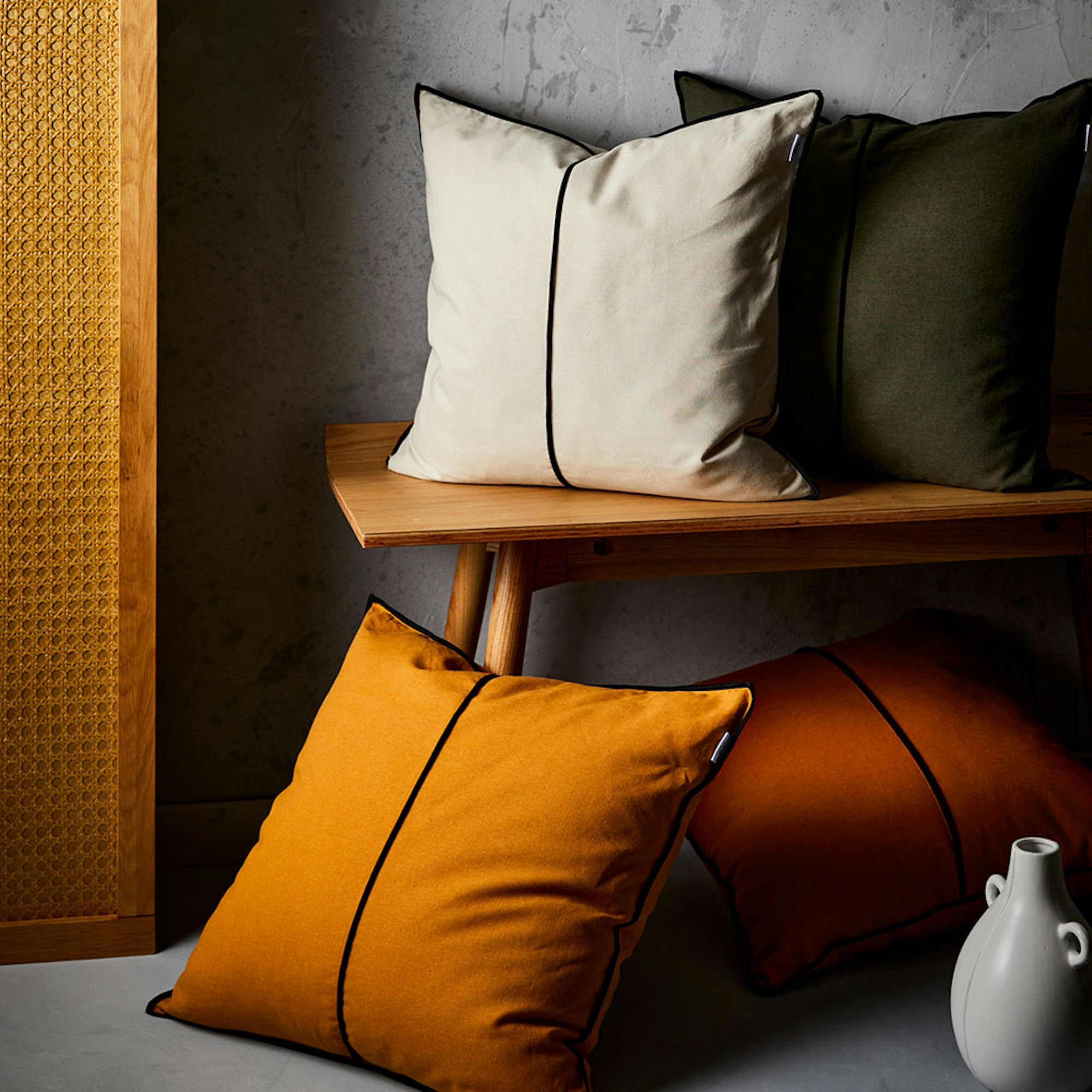 Neale Whitaker SS23 Tooli Cushion. Stacks of cushions in earthy shades with black piping detail