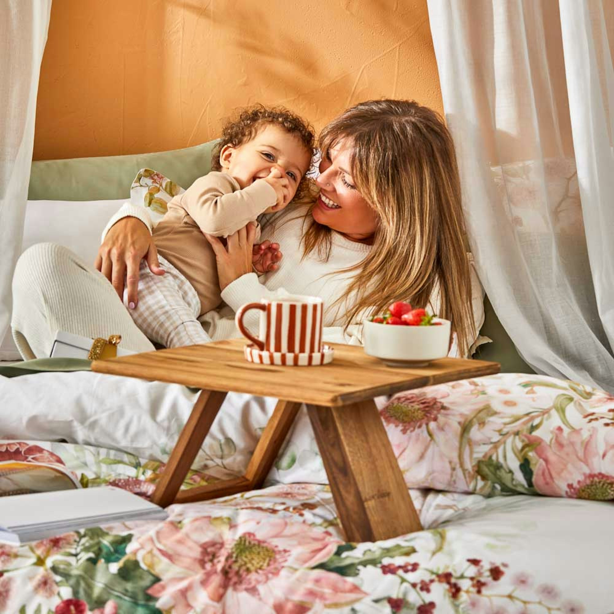 Mother’s Day Gift Guide 2024 | MyHouse blog. Mum sitting on a bed holding her child with a breakfast table and cup of coffee in front of her.