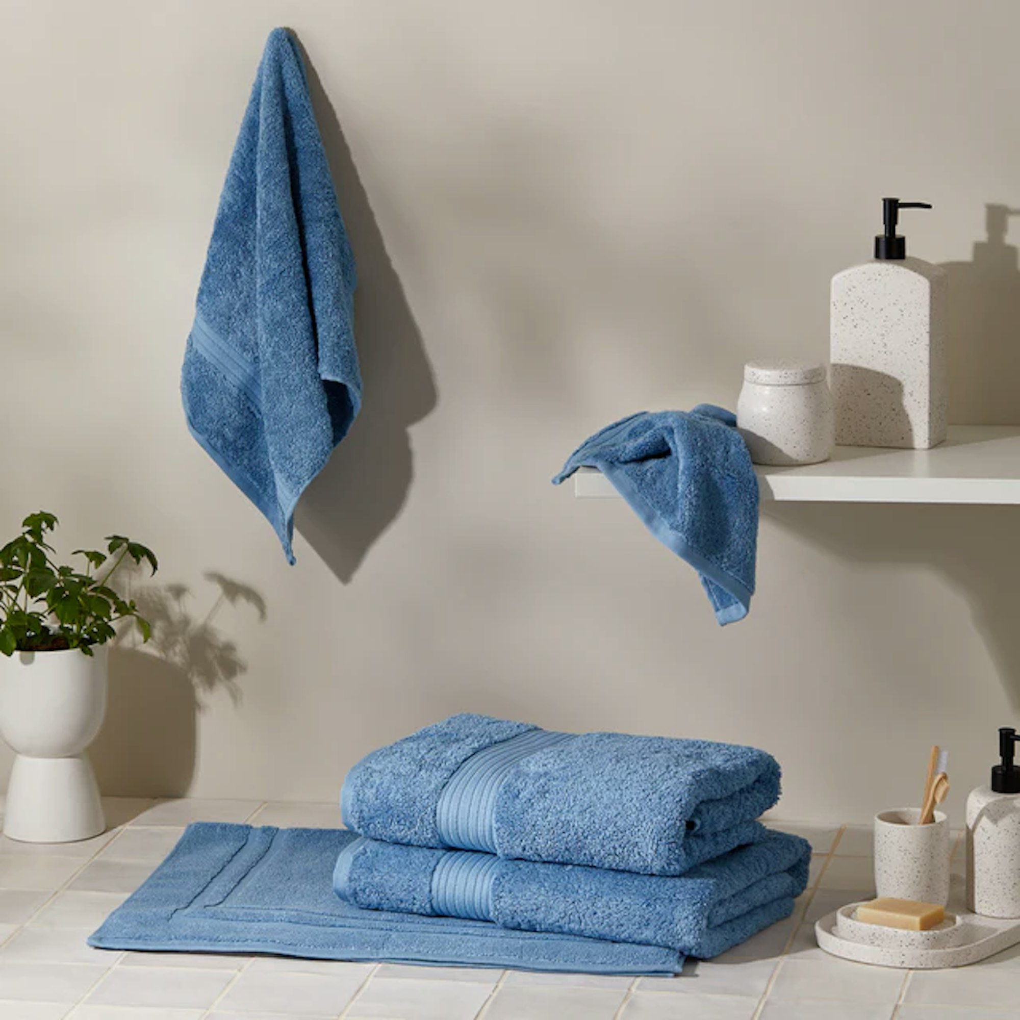 An image of 5 blue towels in a neutral bathroom. One is hanging, one is draped over the corner of a shelf, and two sit folded on top of a bath mat.