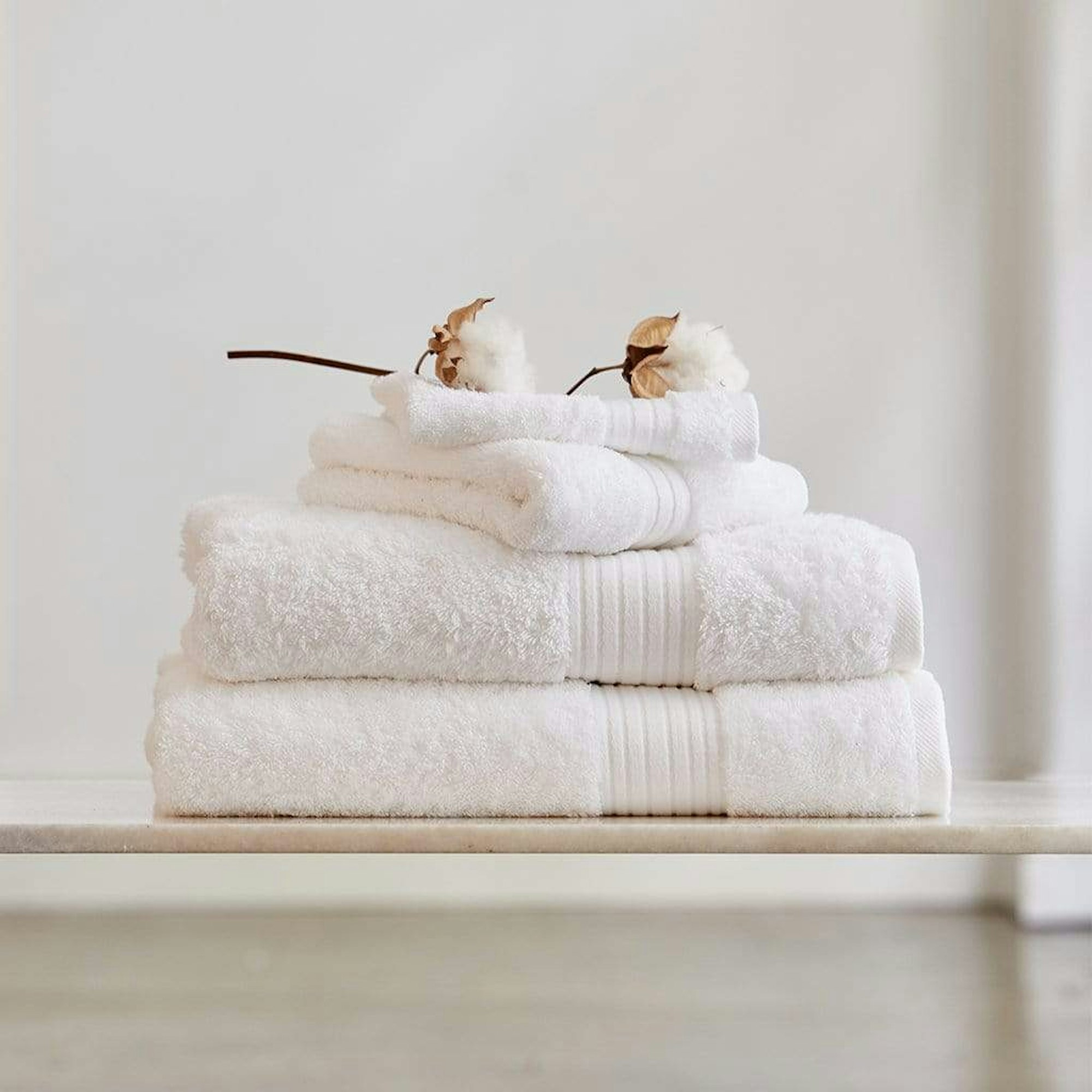 A stack of 4 white towels in front of a neutral background with two cotton flowers on top of the stack