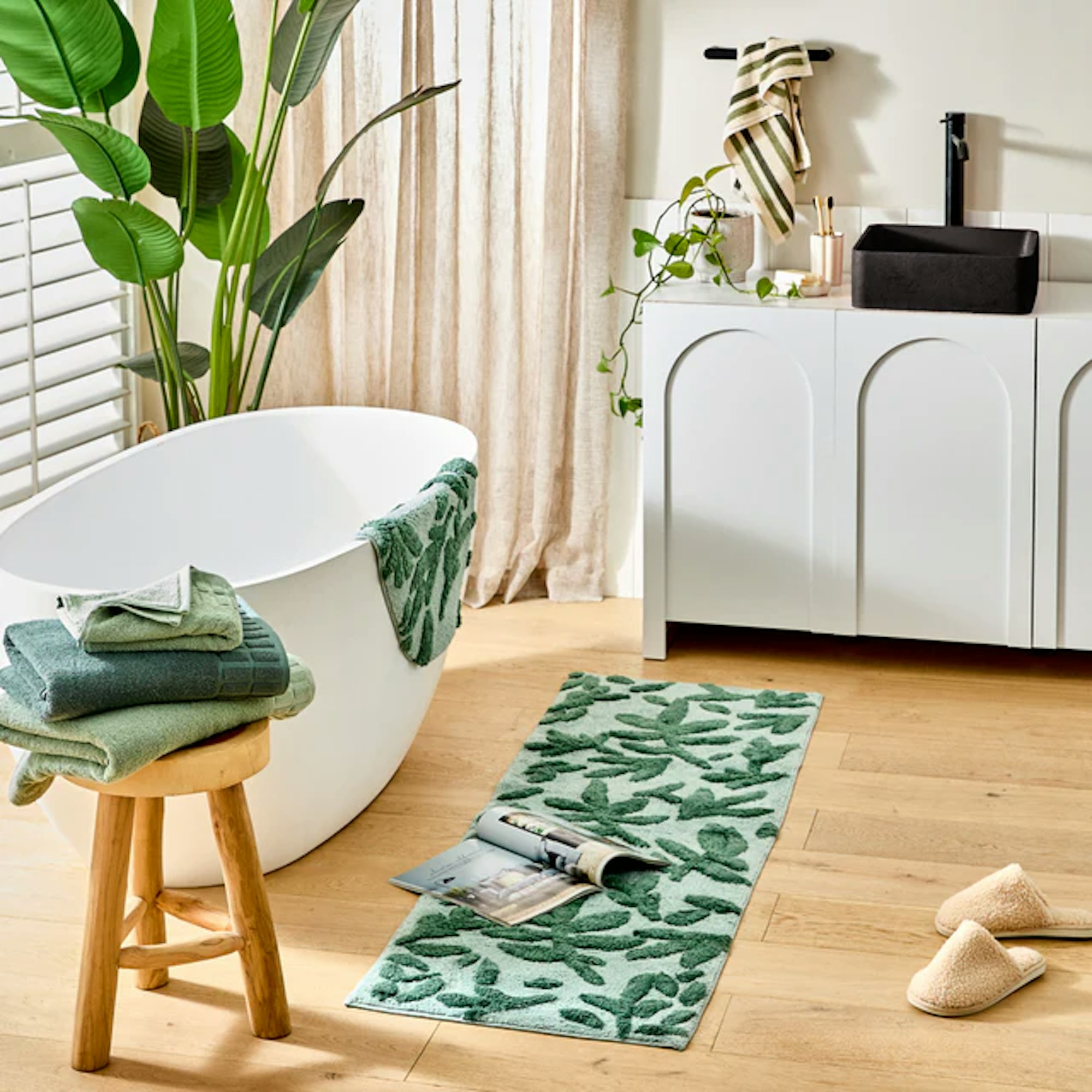 A neutral bathroom with a stack of 3 green towels on a stool in front of a bath with a green leaf pattern bath mat draped over the side and a matching green bath runner on the floor