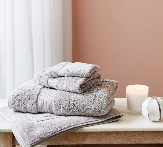 A stack of 3 MyHouse Celene soft grey bath towels and a bath mat on a bathroom bench next to a candle in front of a brown wall