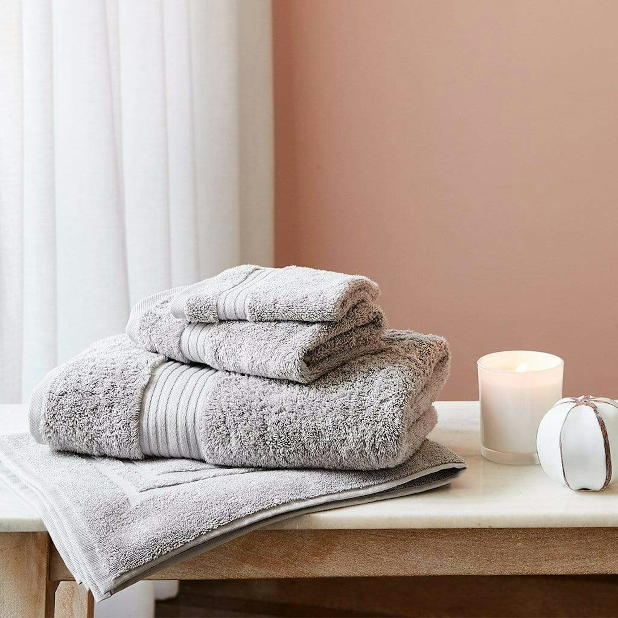 A stack of 3 soft grey bath towels on a bathroom bench next to a candle in front of a brown wall