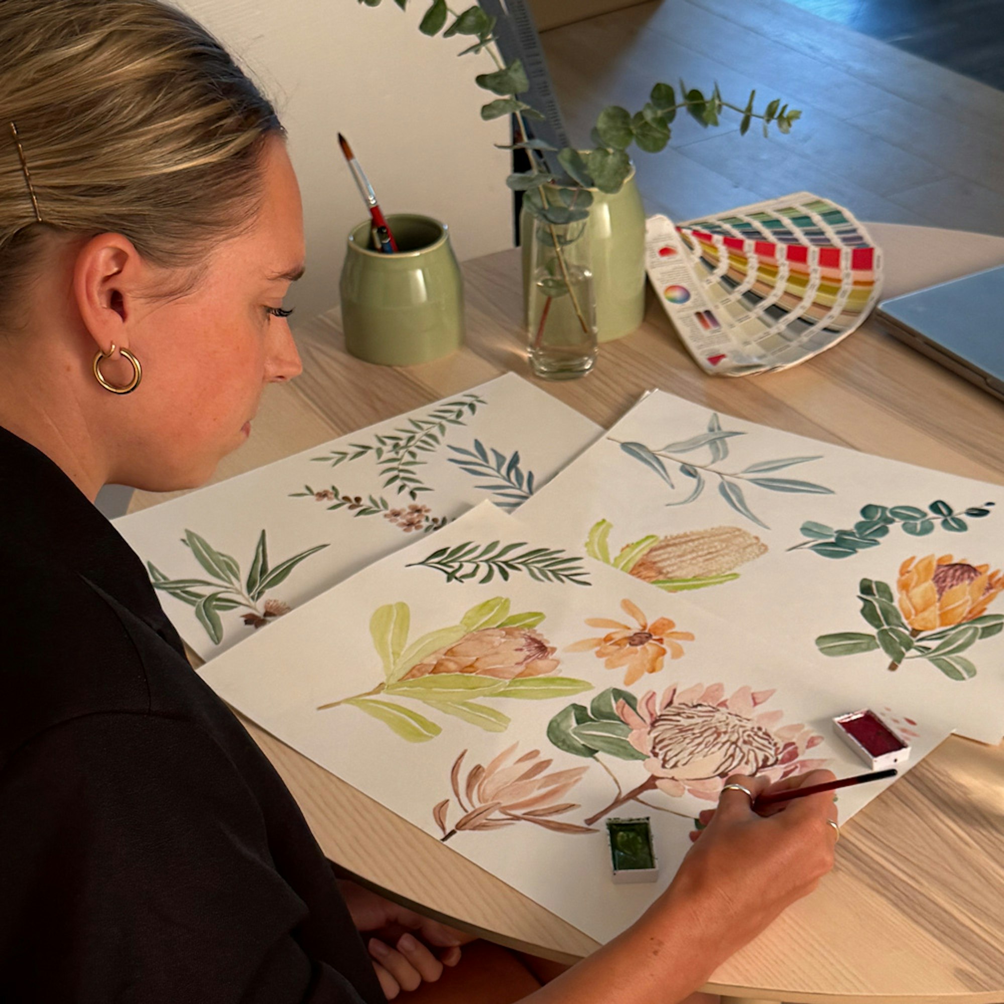 Woman wearing black sitting at a wooden desk painting floral illustrations