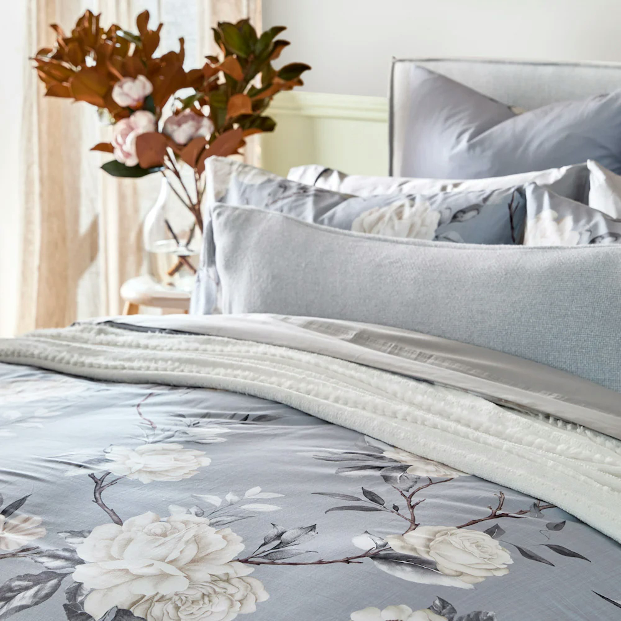 Close up of a light blue quilt cover and matching pillowcases with a white floral design and autumnal flowers in a clear vase in the background