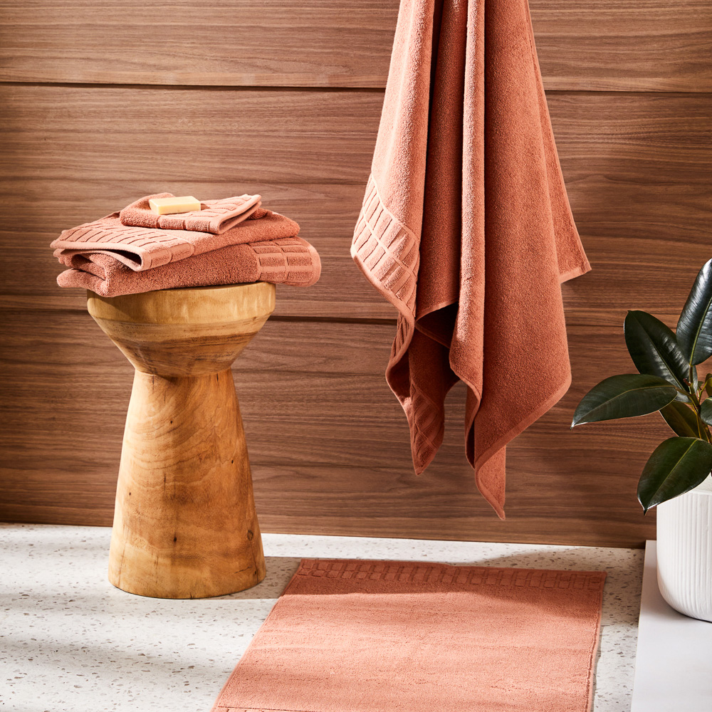 A stack of pink towels on a wooden stool with one towel hanging on a hook in front of a brown wall