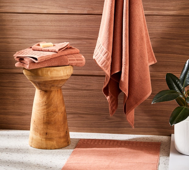 A stack of pink towels on a wooden stool with one towel hanging on a hook in front of a brown wall