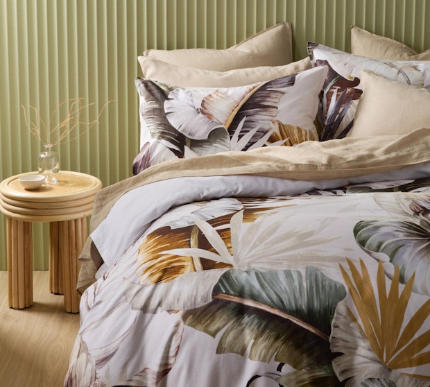A white quilt cove with matching pillowcases with a leaf design in front of a green wall and next to a wooden beside table.