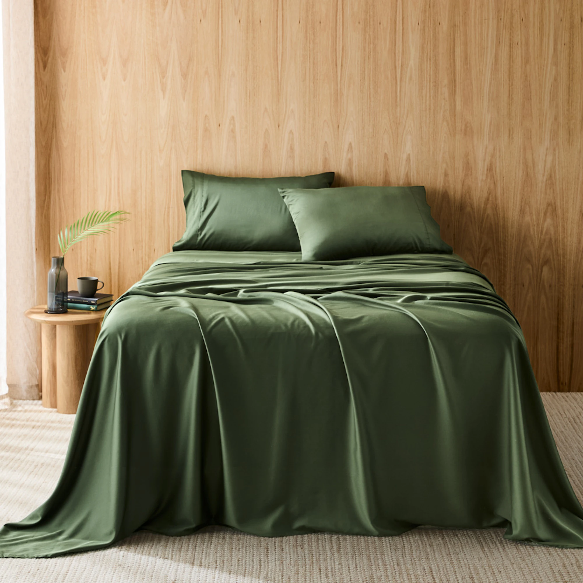 A moss green queen bed sheet set against a bamboo coloured wall. Featuring a draped flat sheet, a fitted sheet, and two pillowcases.