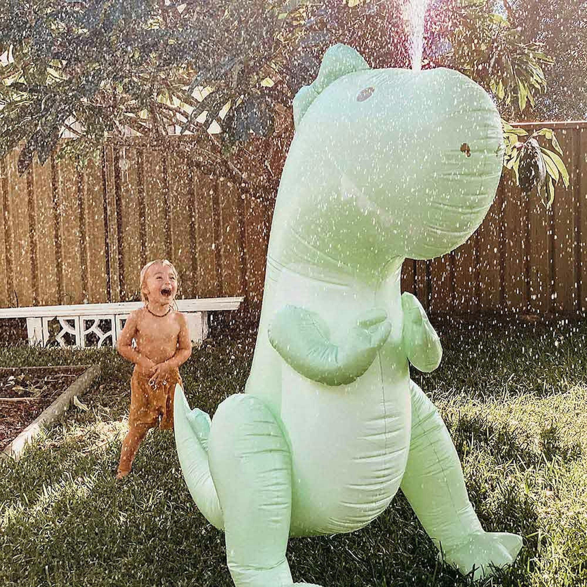 Child playing outside with giant green dinosaur sprinkler.