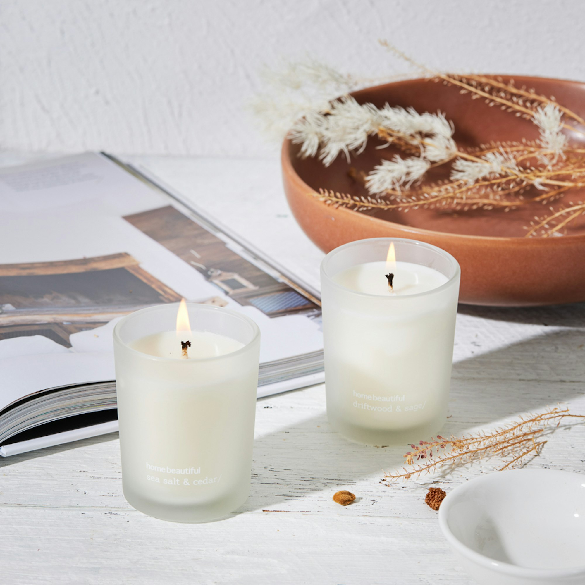 Home Beautiful Scented Candles Collection. Lit candles with open book and rust bowl.