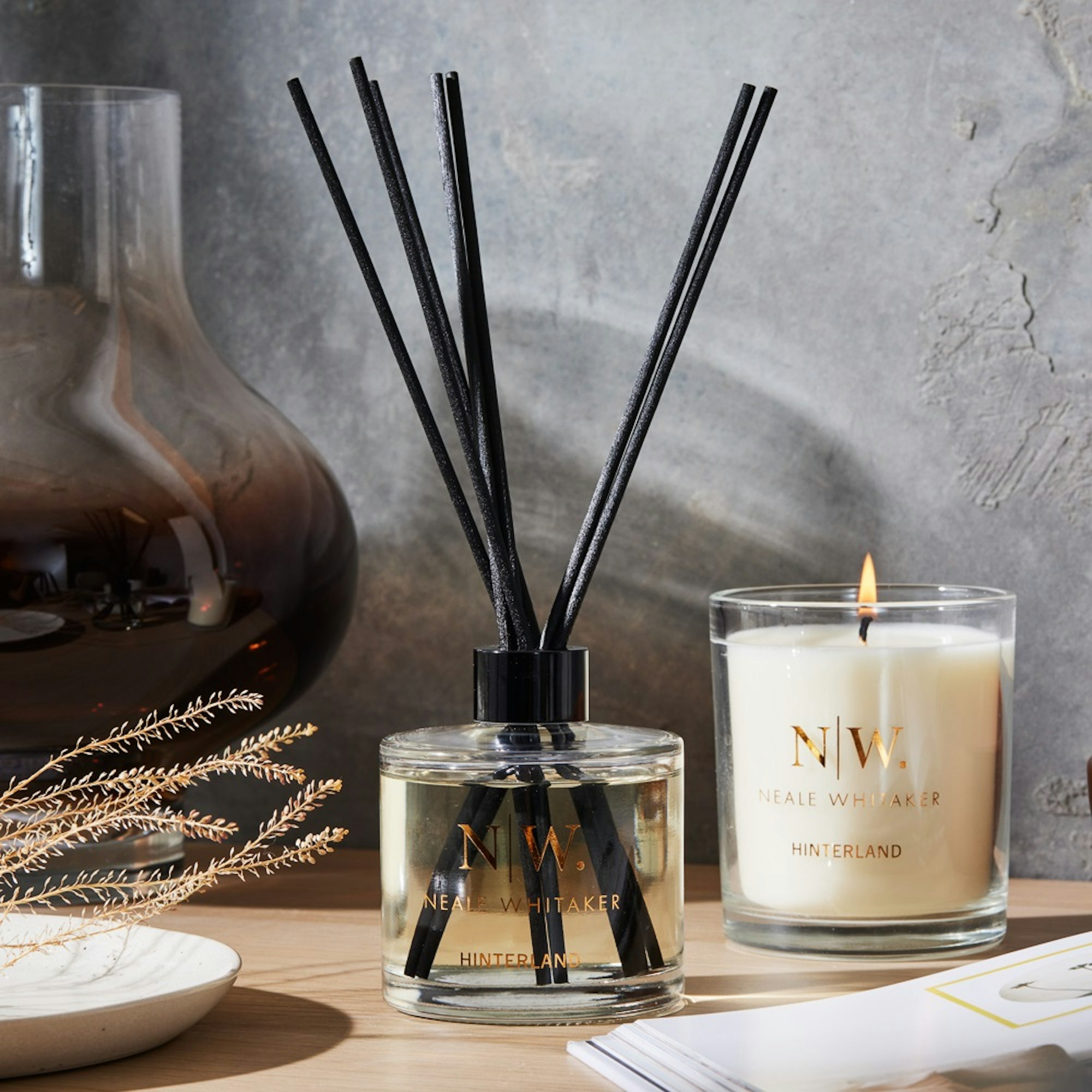 Neale Whitaker scented candle and diffuser. Dark and moody setting with lit candle and textured wall.