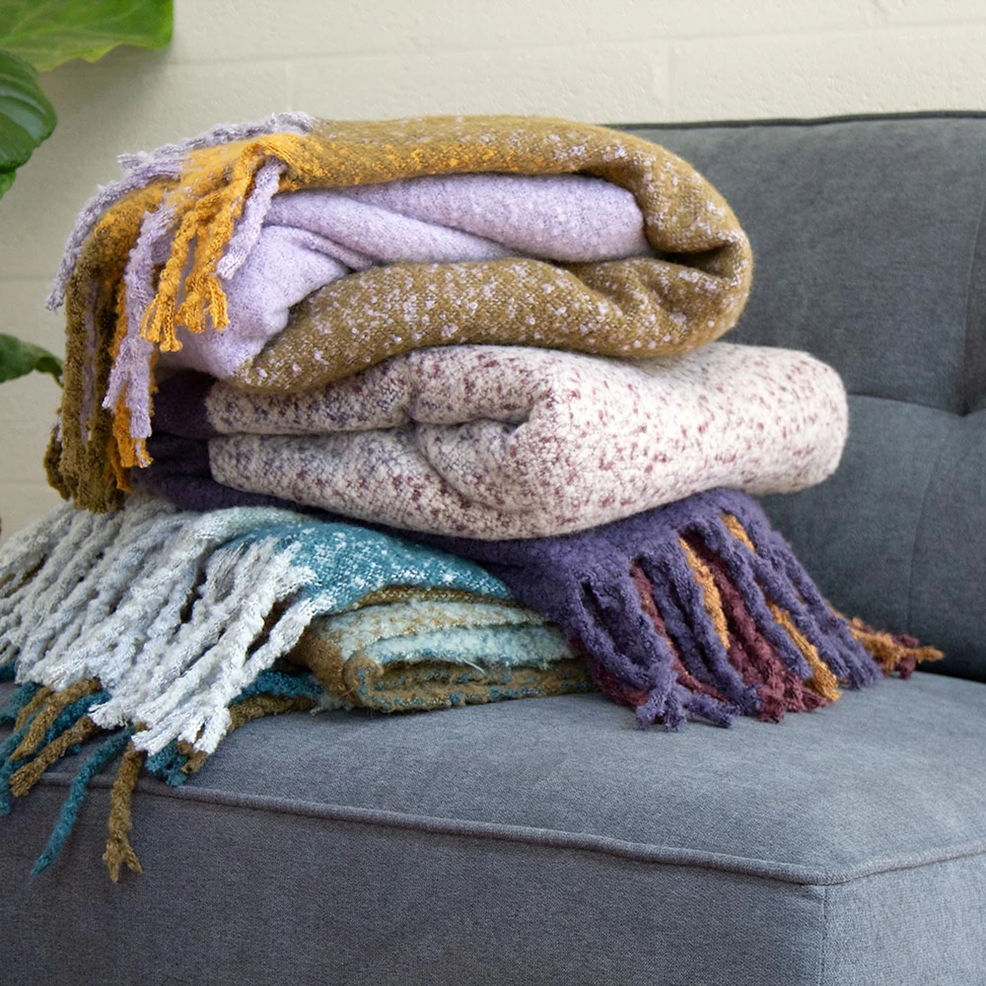 Bambury throws in assorted colors folded on a grey sofa, showcasing soft textured fabric and fringed edges