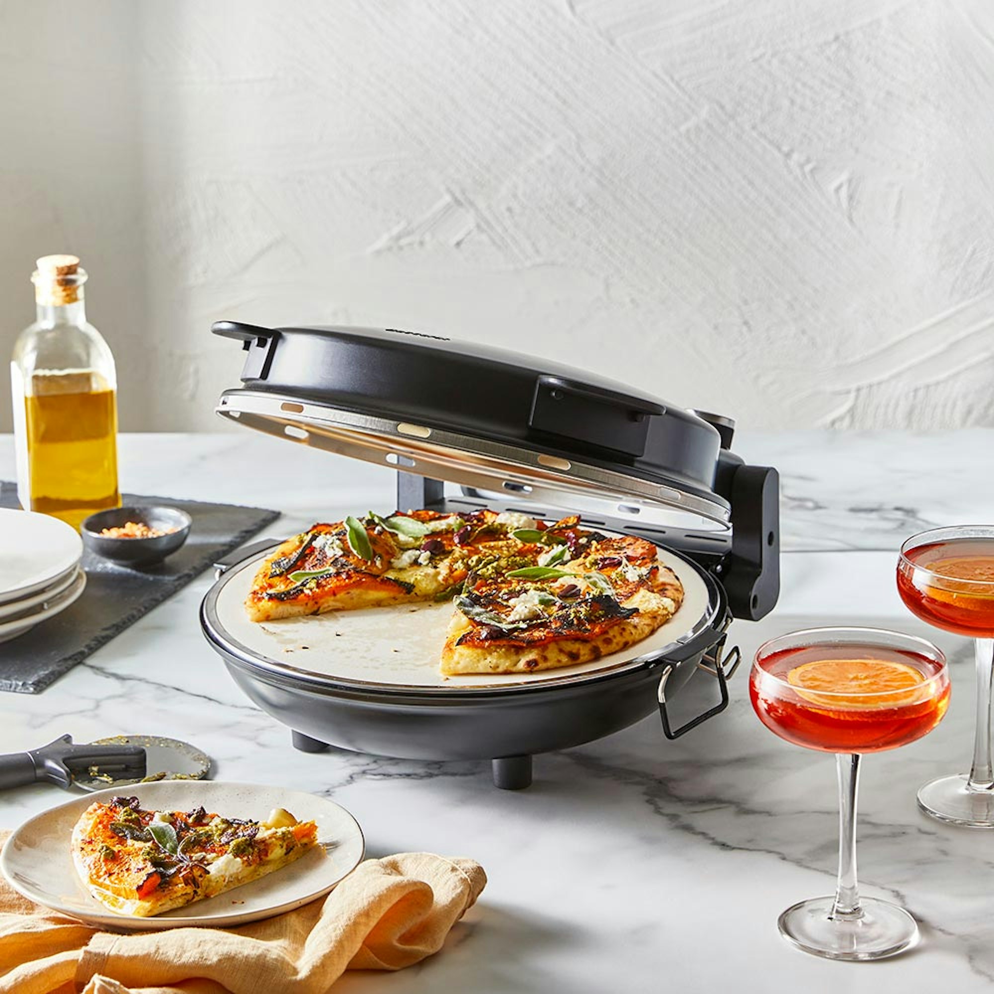 Baccarat the Gourmet Slice Pizza Oven Black on a bench with pizza and 2 drinks