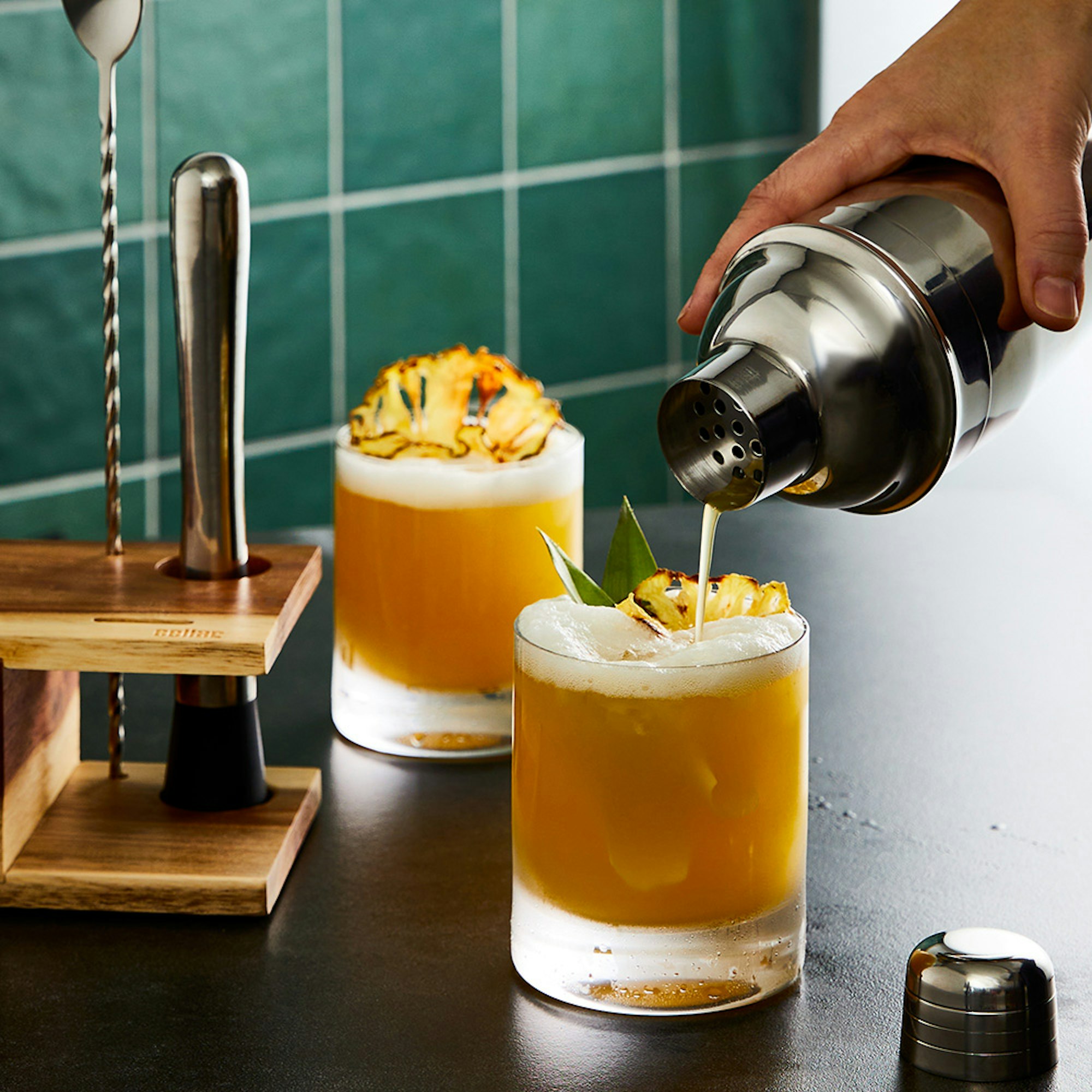 Father's day gift guide. 7 Piece bar tool set. man poring two cocktails into tumblers with pineapple garnish.