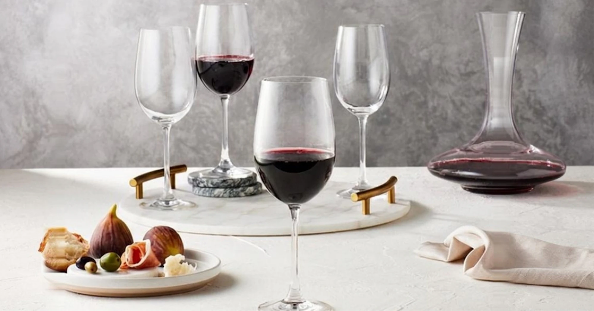 Four red wine glasses and a decanter set out stylishly on a white table with a plate of figs
