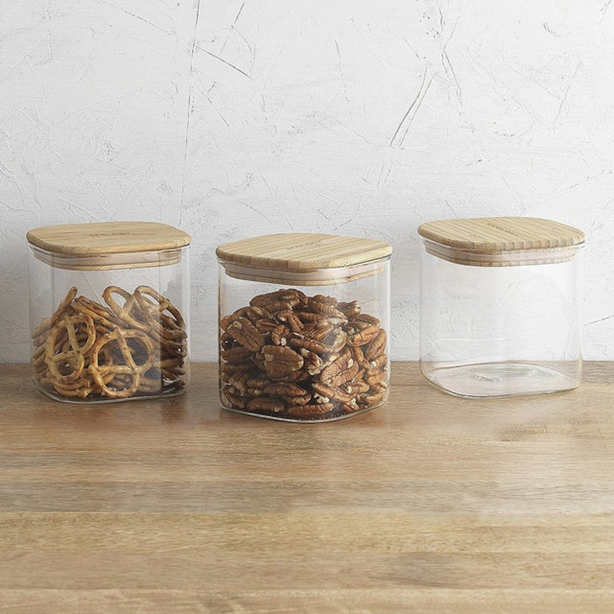 Set of three clear storage canisters with bamboo lids, containing pretzels and pecans, displayed on a wooden surface against a textured white wall