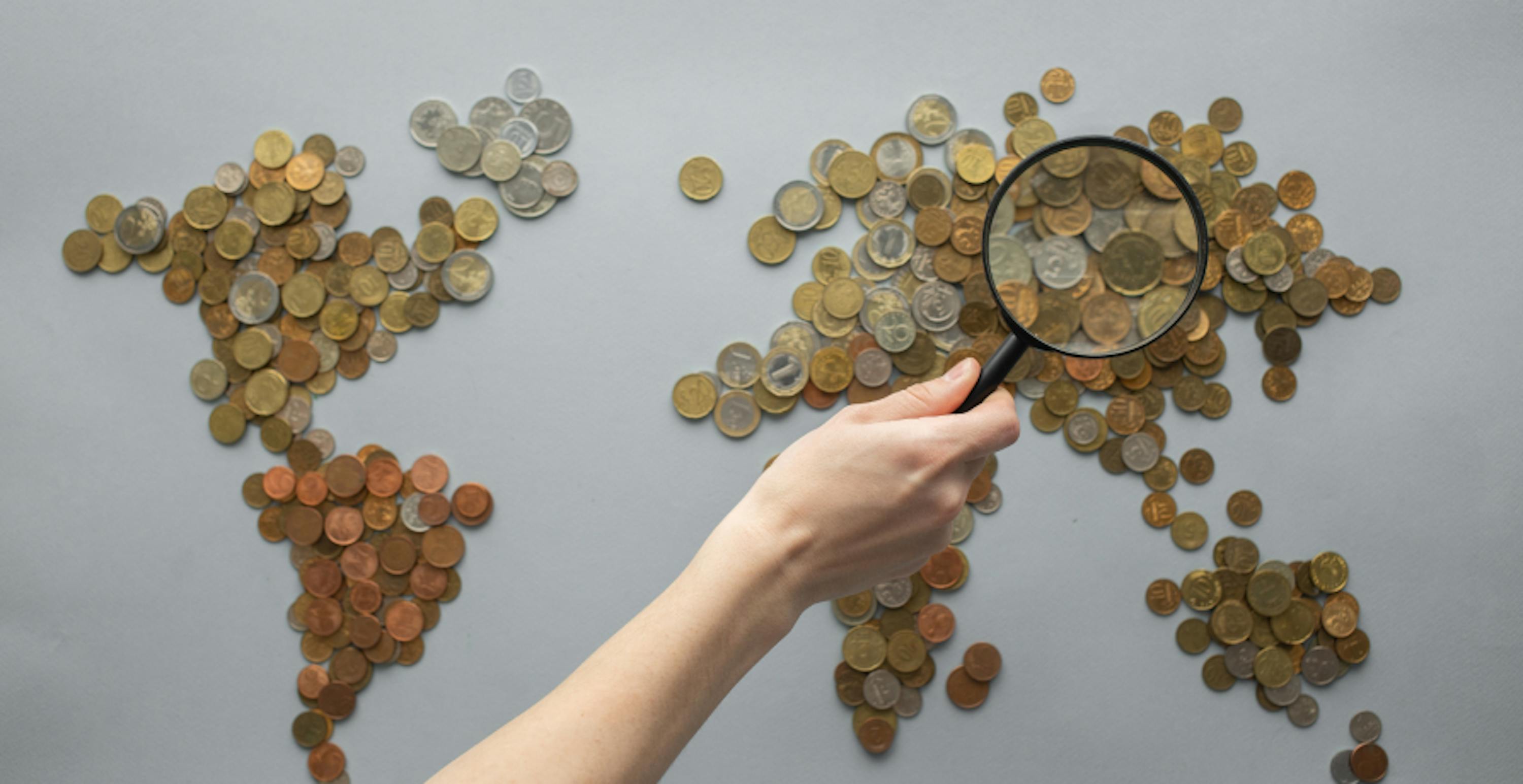 person holding magnifying glass looking at map of the world made from money