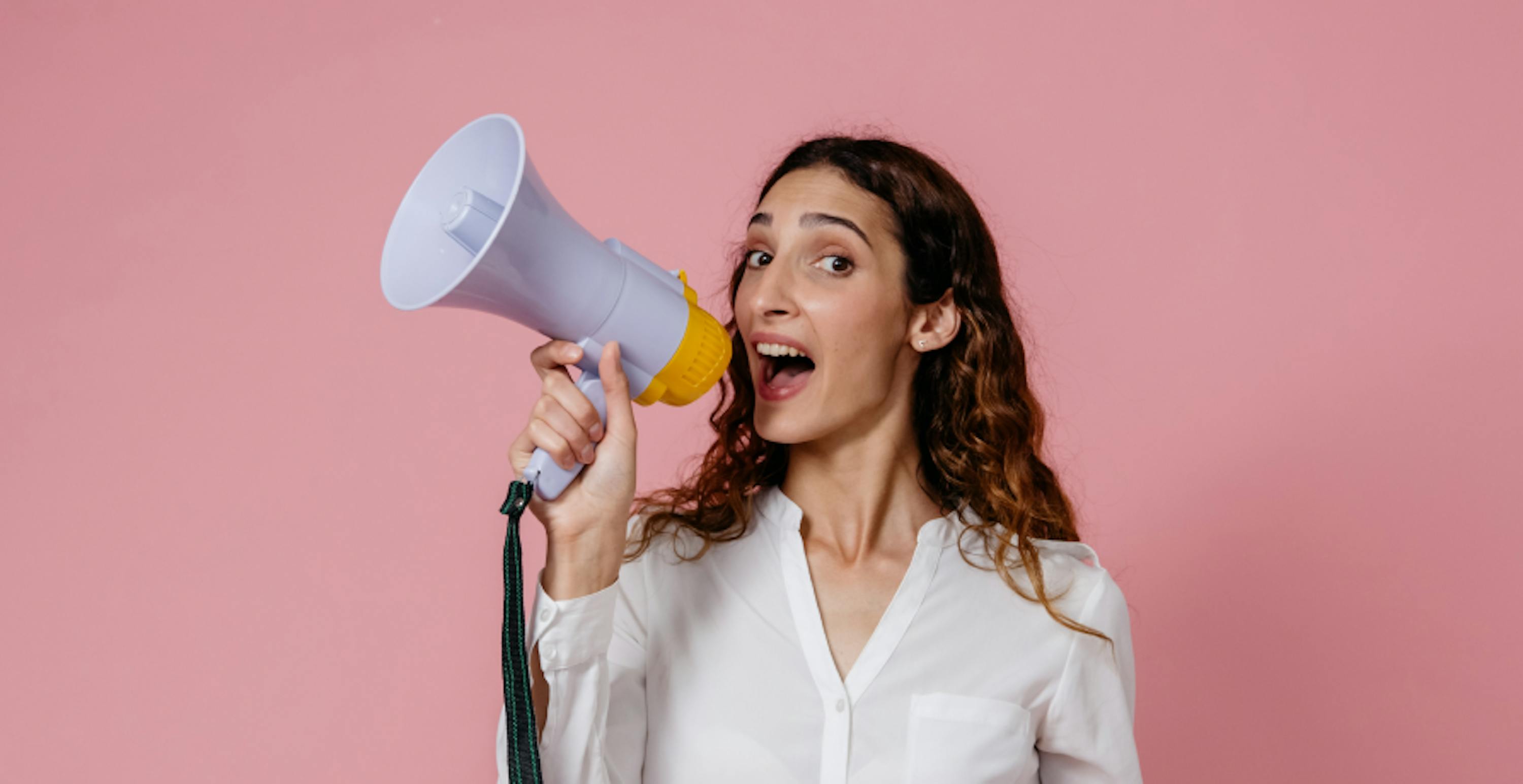 femme debout devant un mur rose criant dans un mégaphone 