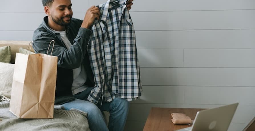 man taking clothes he bought of out bag with laptop open