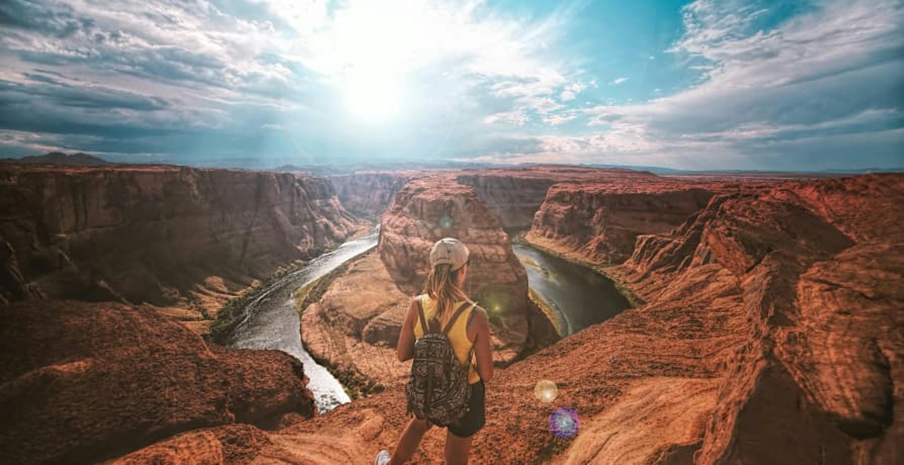 person yellow shirt shorts blonde hair wearing backpack standing over view of grand canyon blue sky sunny