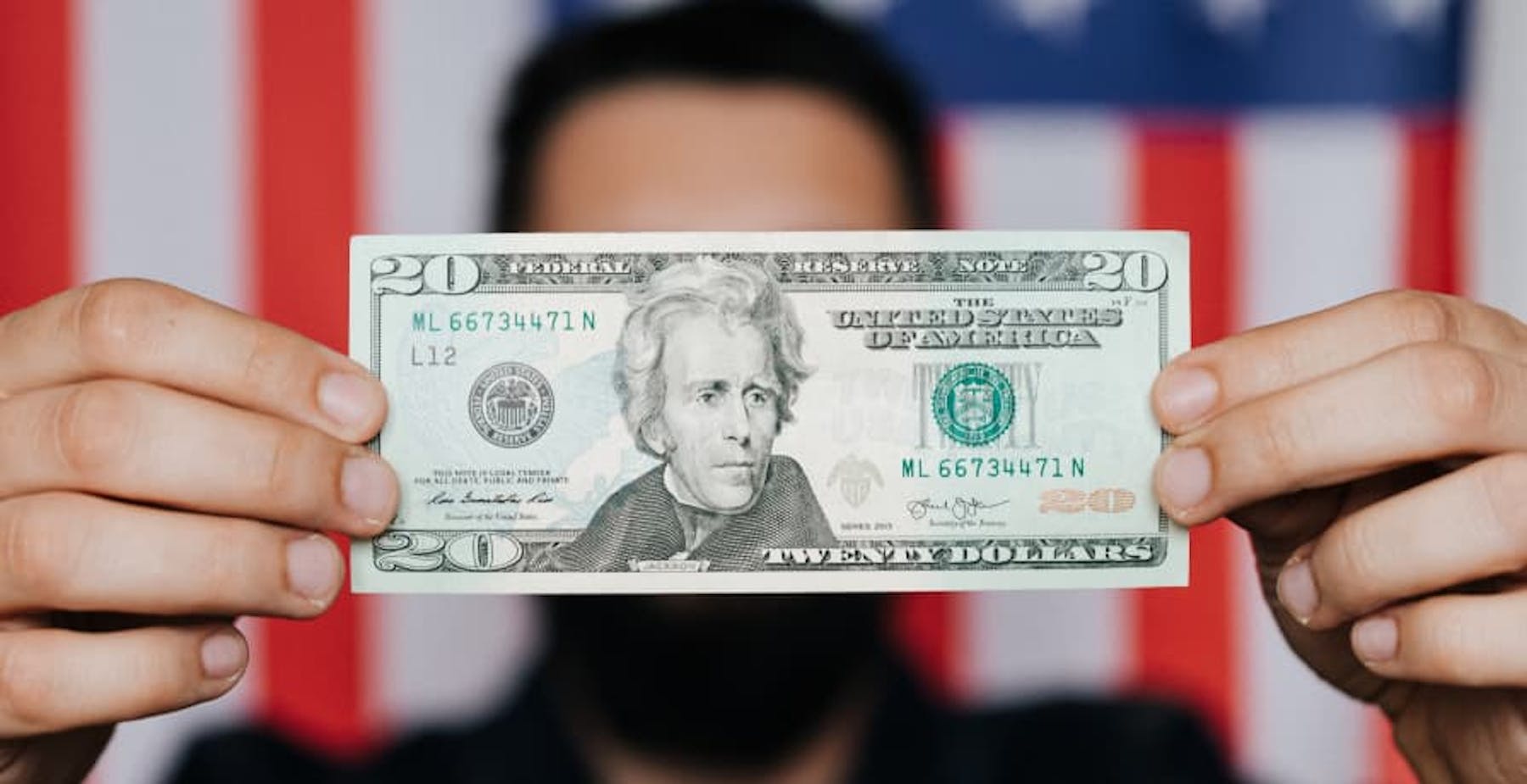person holding up single USD $20 bill in front of US flag