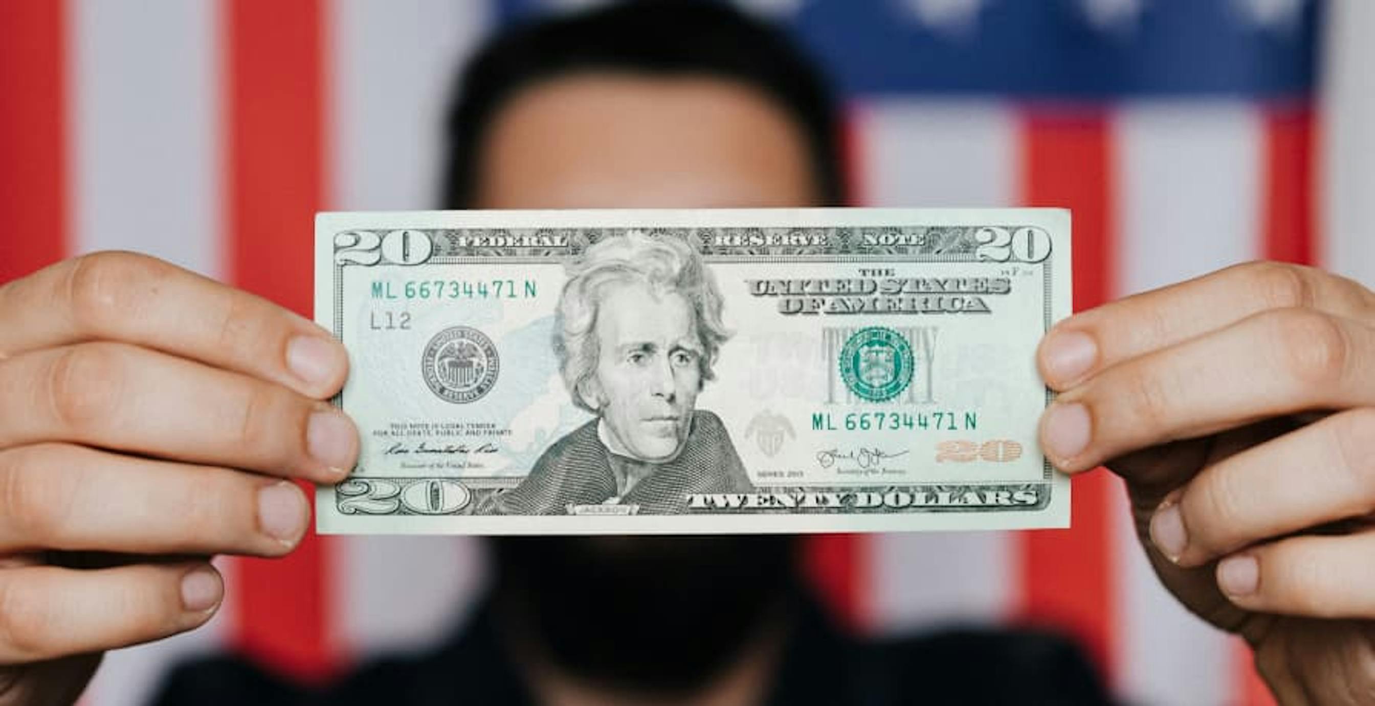 person holding up single USD $20 bill in front of US flag