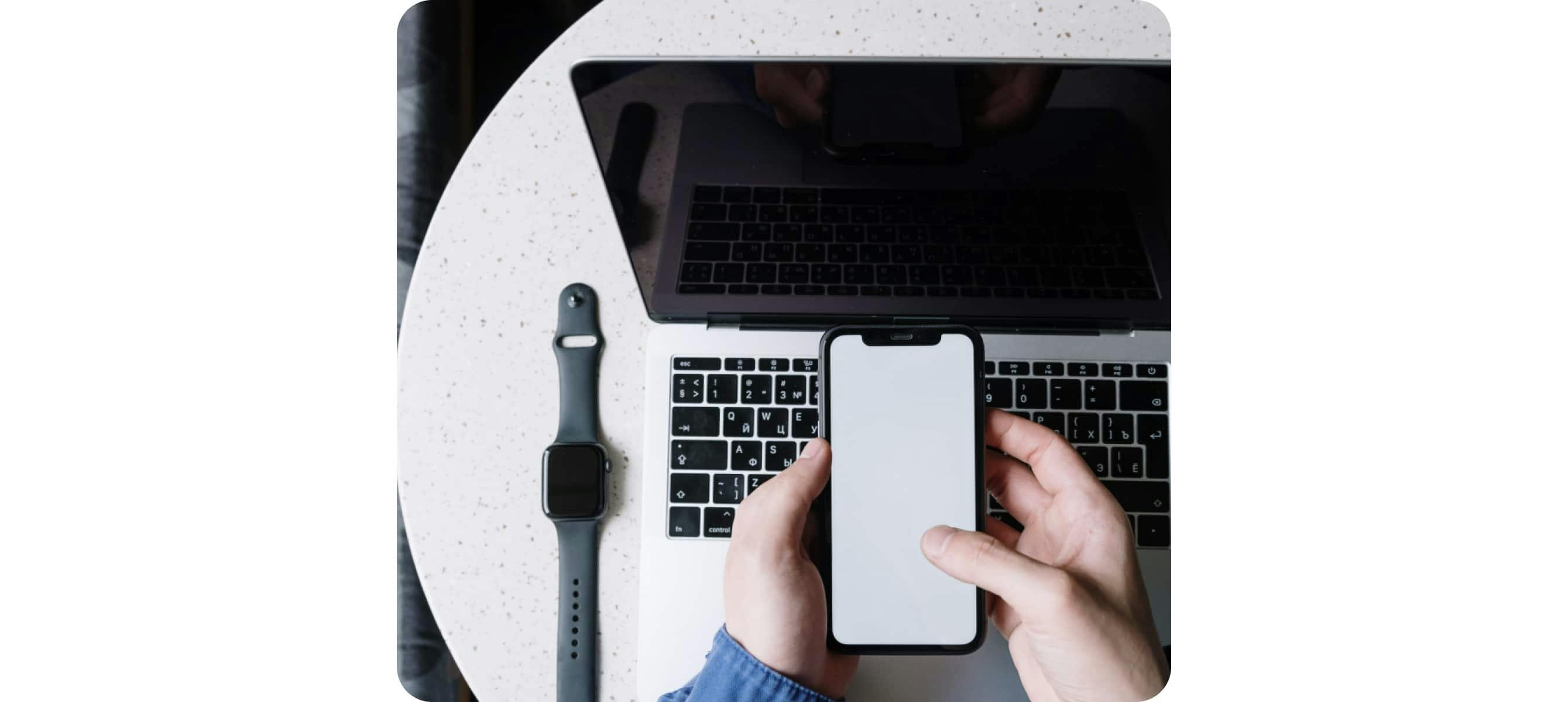 une personne tenant un téléphone avec un ordinateur portable et une smartwatch sur la table