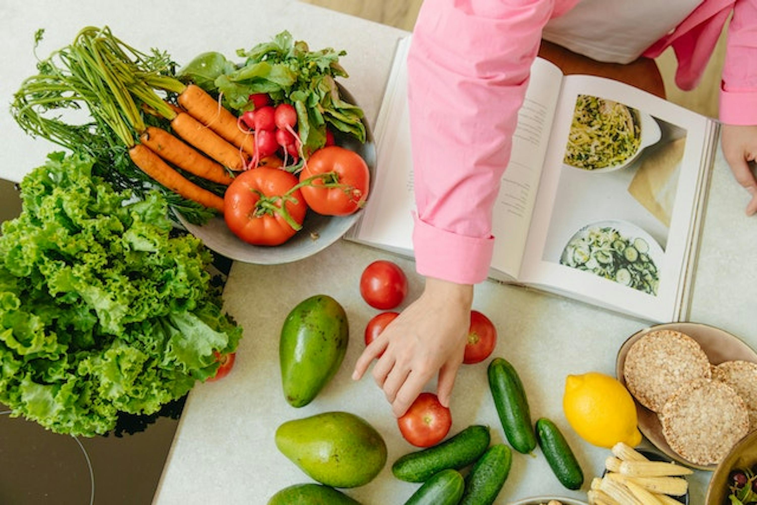 Personne prenant une tomate sur une table remplie de légumes, tout en lisant un livre de recettes