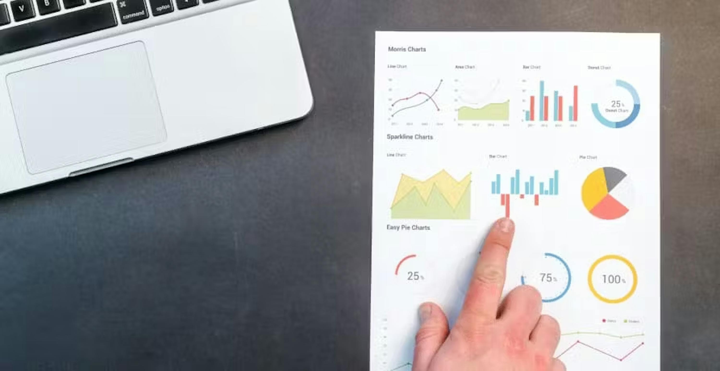 person pointing to graphs and data on piece of paper with laptop in the background