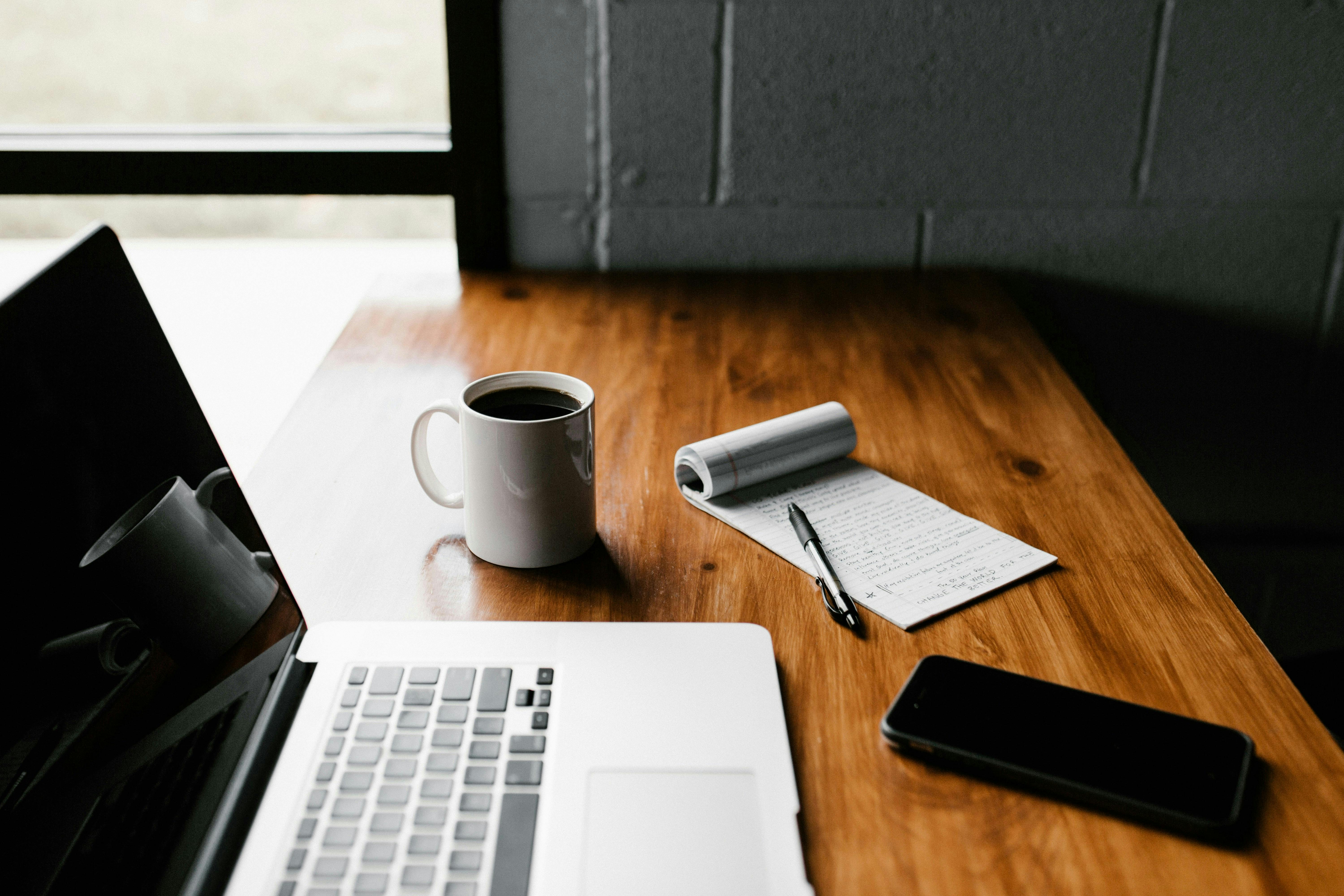 desk with a laptop, smartphone, coffee mug and notepad