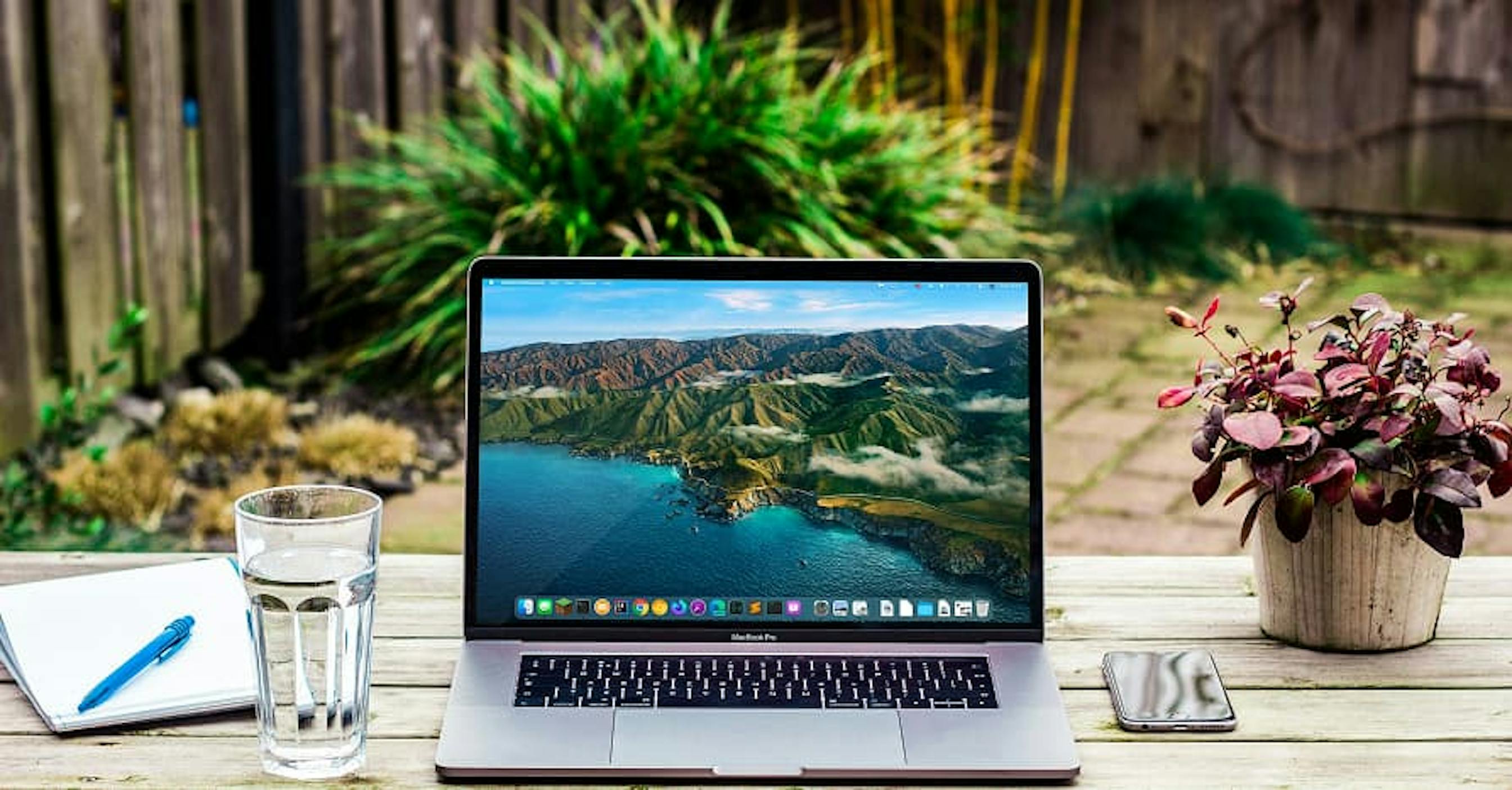 laptop open to cliff-sea-side view with flowers glass of water and notepad