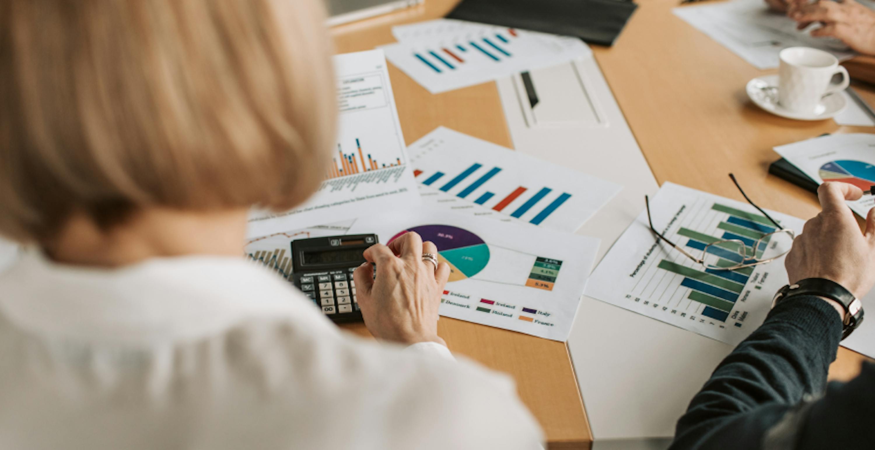 Une femme de dos assise devant une table recouverte de documents montrant des statistiques en train de taper sur une calculatrice