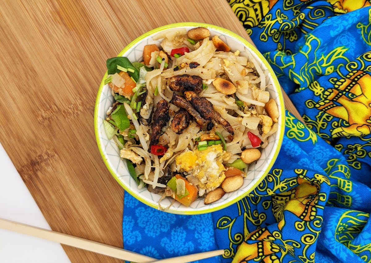 Pad thai topped with marinated grasshoppers in a small bowl and decorations