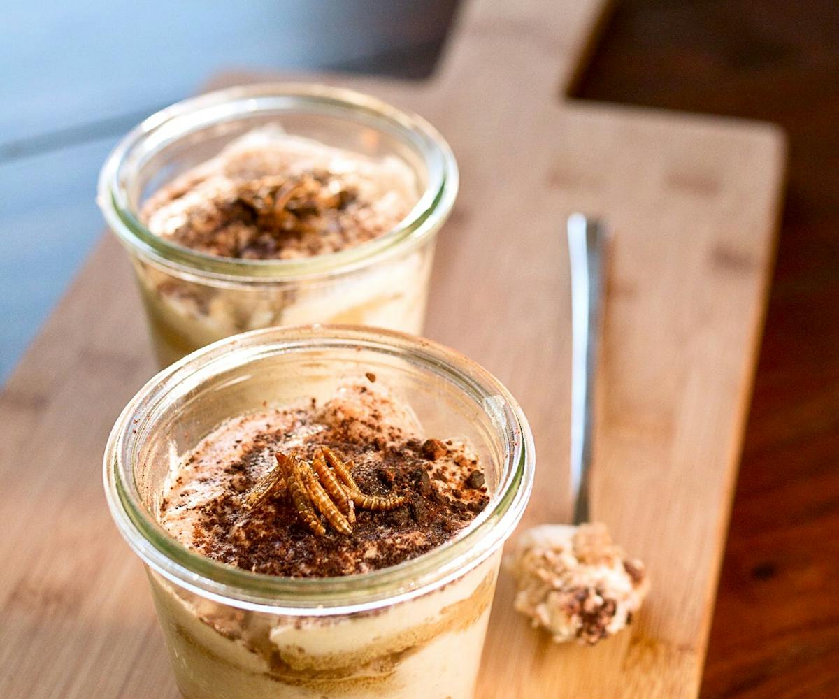Cup and spoon of tiramisu topped with mealworms on a cutting board