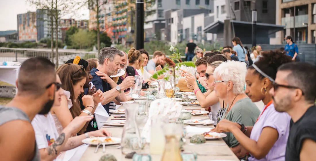 Group of people eating lunch together on long table inside Greenwich Peninsula